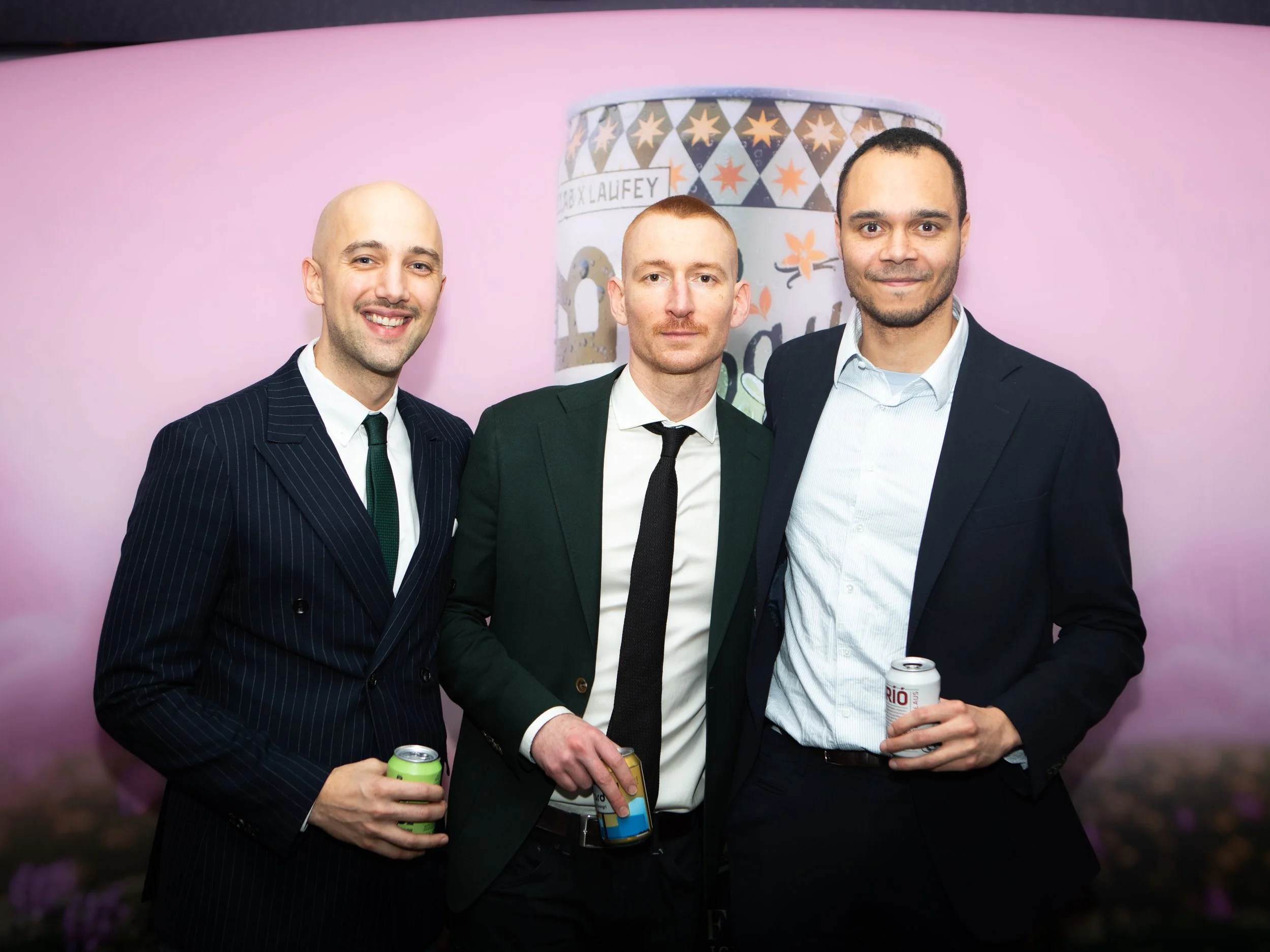 Three men in suits standing together, smiling, holding canned drinks, with a pink background and a decorative large can with stars and text behind them.