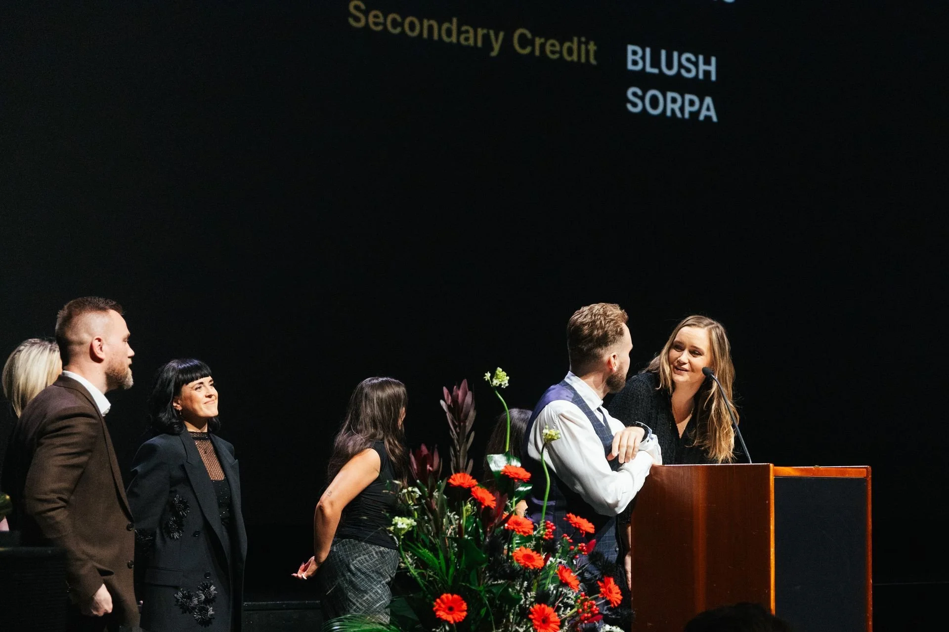 Group of people standing on stage at a formal event, with a woman speaking into a microphone at a wooden podium, surrounded by floral arrangements, under a large screen displaying text related to academic credits.