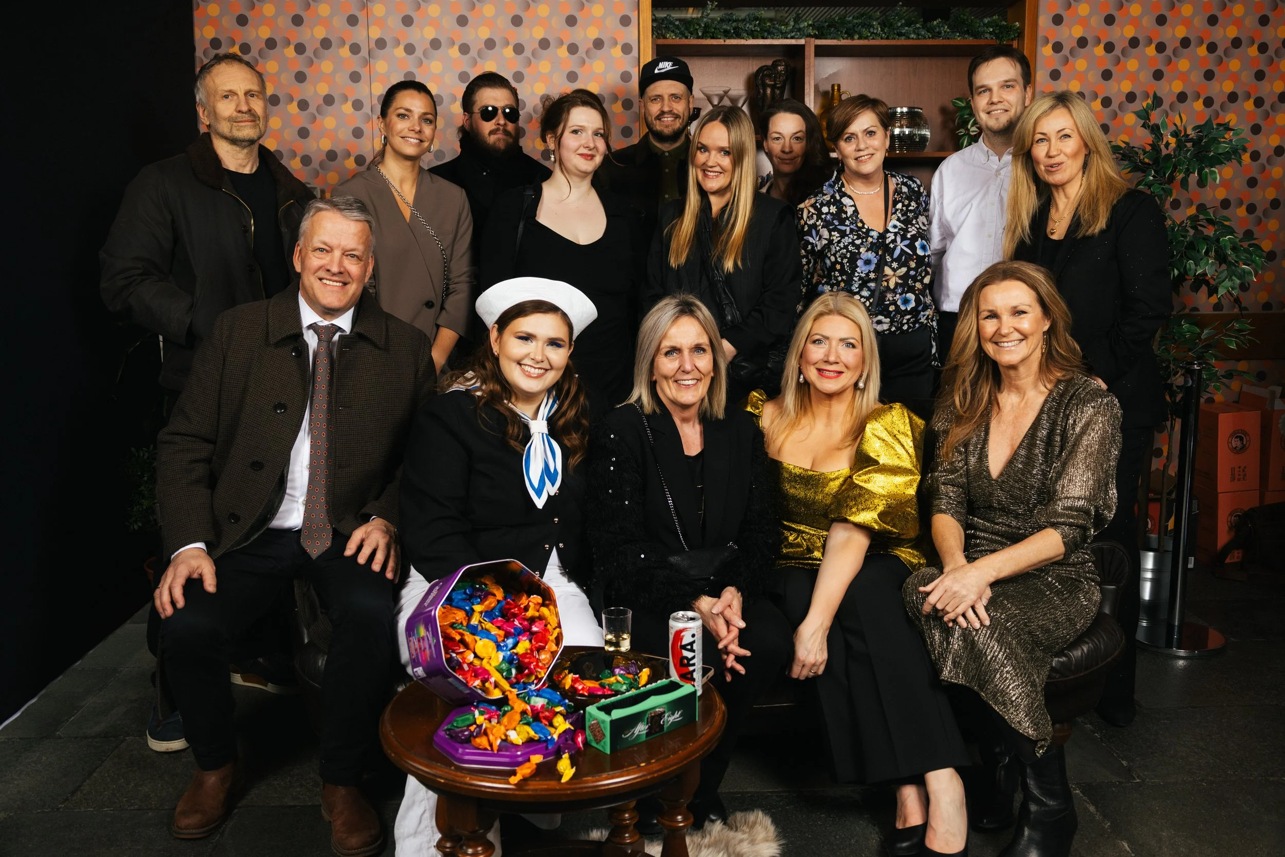 Group of 13 people in a room, some sitting and some standing, celebrating with a table that has a purple box of colorful chocolates, a can of soda, and a glass. The background has colorful circular patterned wallpaper and shelves with decorations and