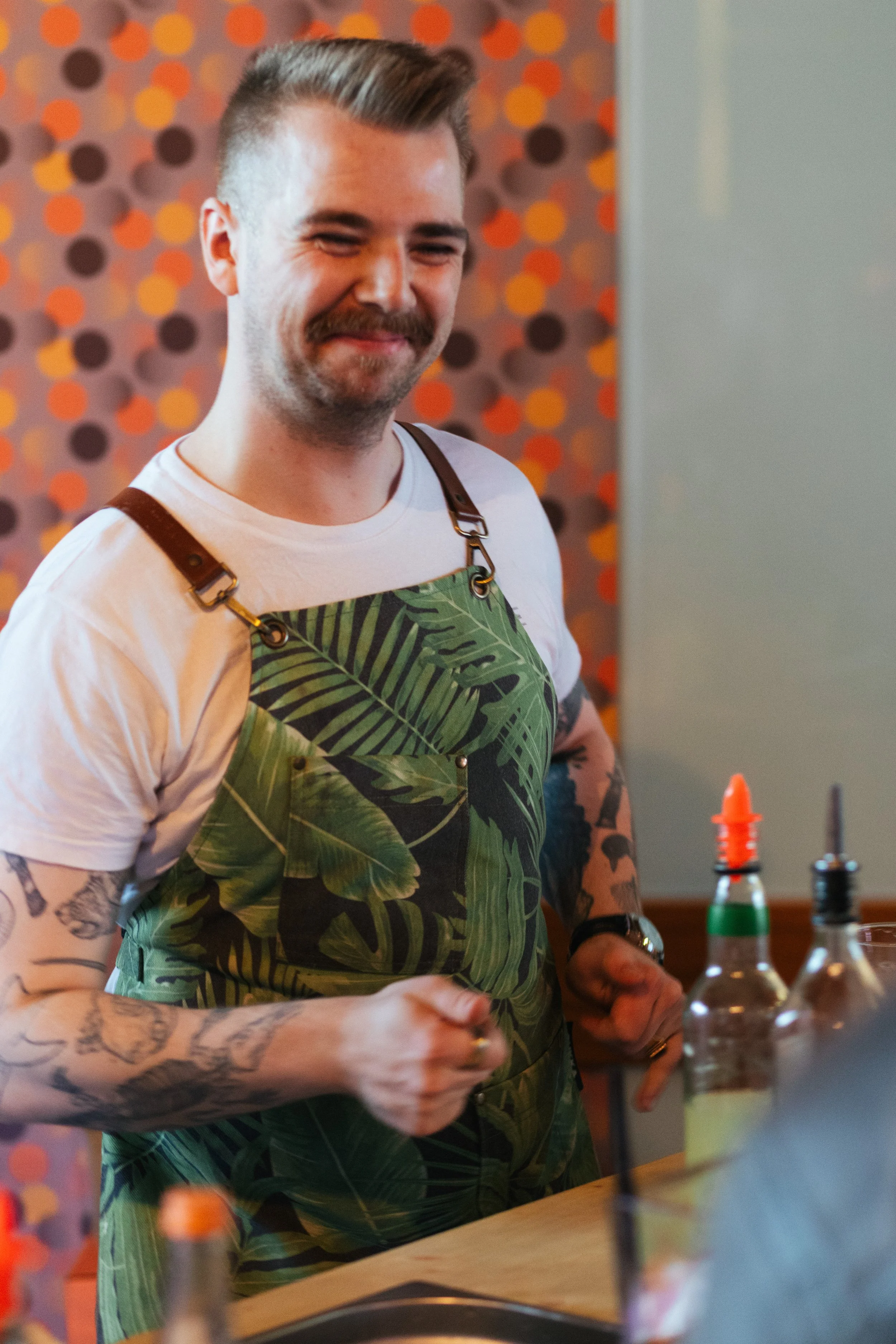A smiling man with a mustache and tattoos on his arms wearing a white shirt and green tropical print apron, standing in a restaurant with a polka dot patterned wall in the background.