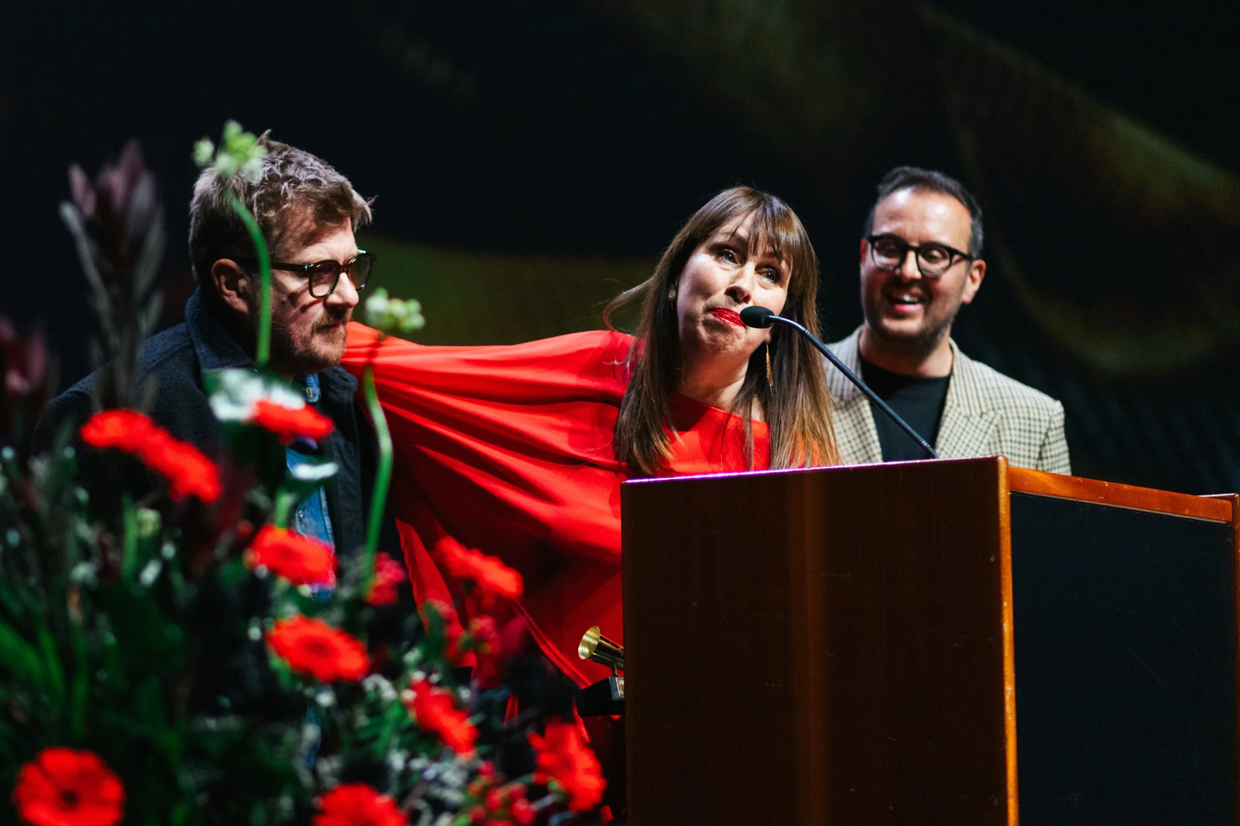 Three people speaking at a podium during a public event, with red flowers in the foreground.