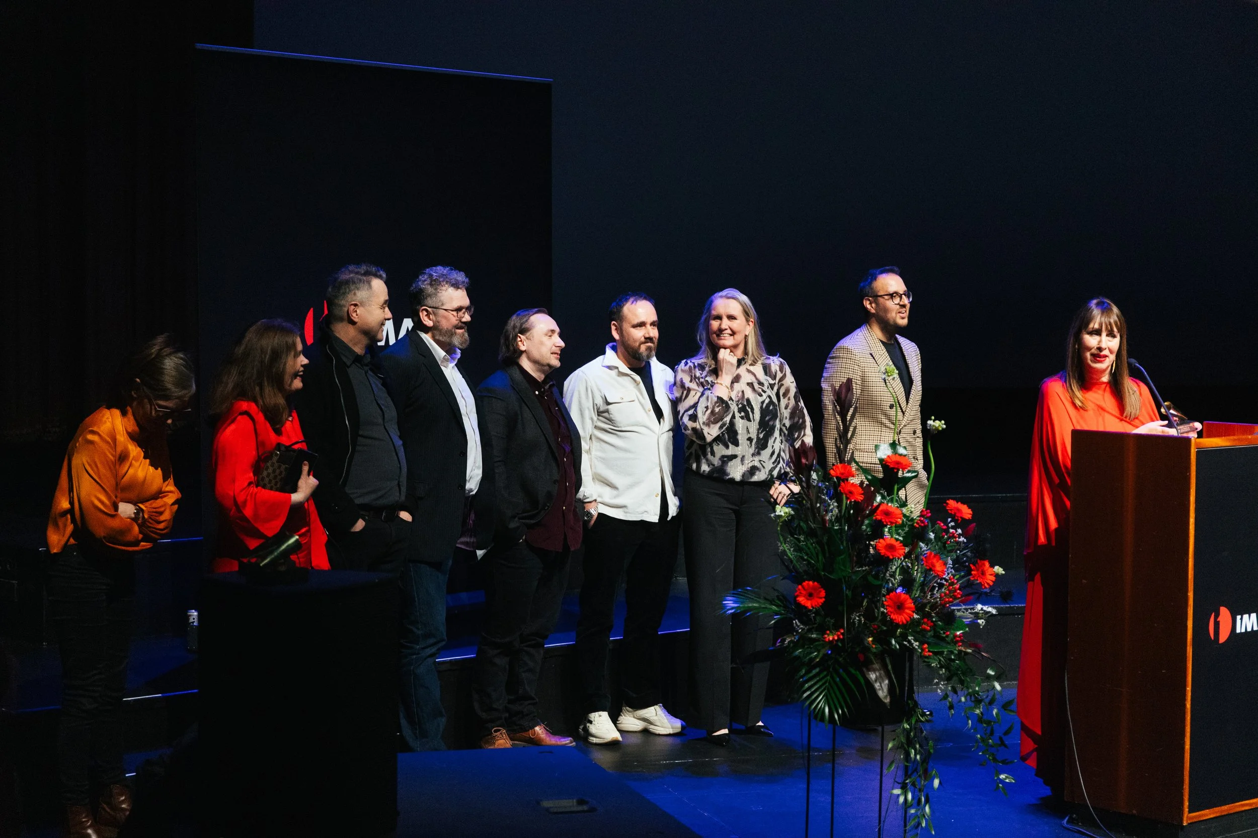 A group of nine people standing on stage during an event, with a woman at a podium speaking into a microphone. The stage has a black backdrop with some lights and floral arrangements in front of the podium.