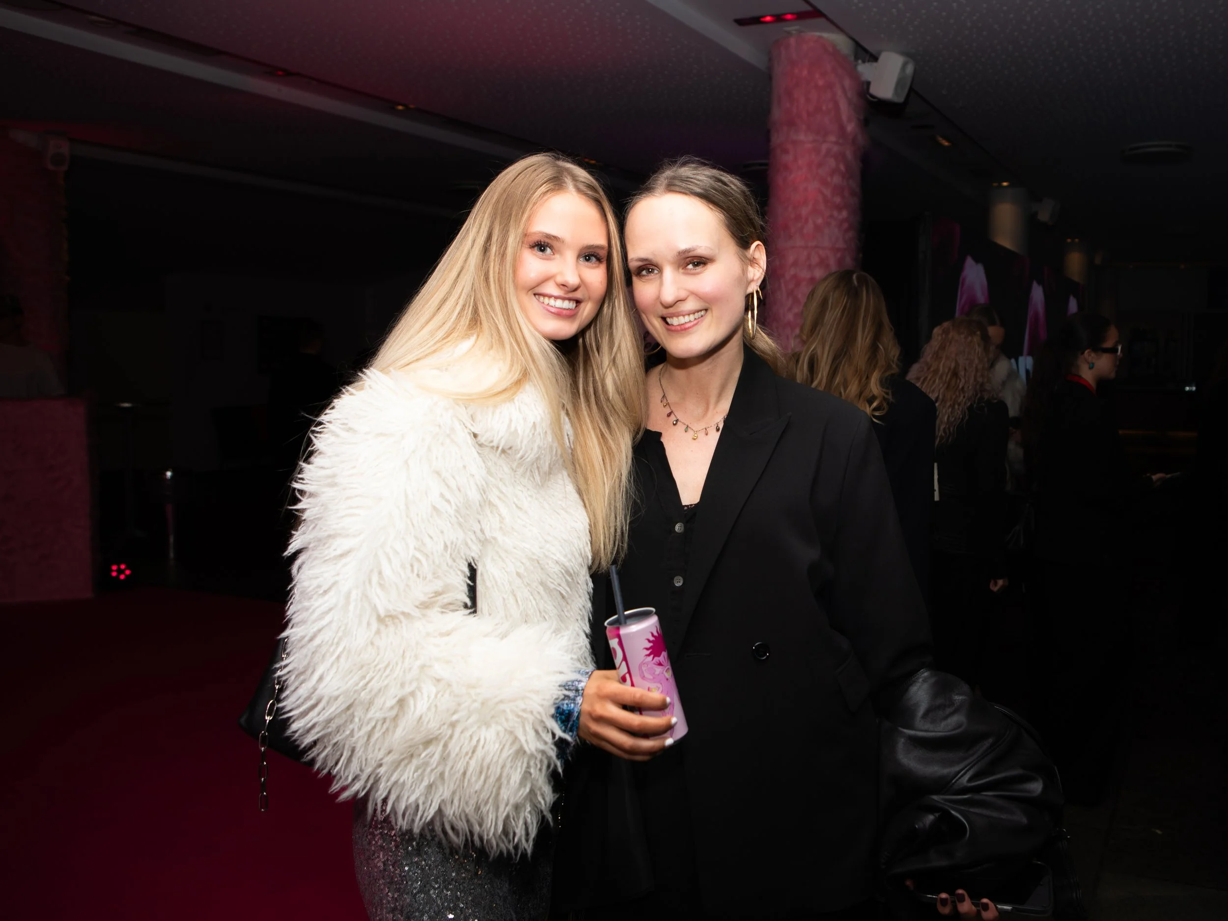 Two women smiling and posing together in a nightclub or event setting. One woman has long blonde hair and is wearing a white fuzzy coat, the other has brown hair, earrings, and is dressed in black, holding a pink and white drink.
