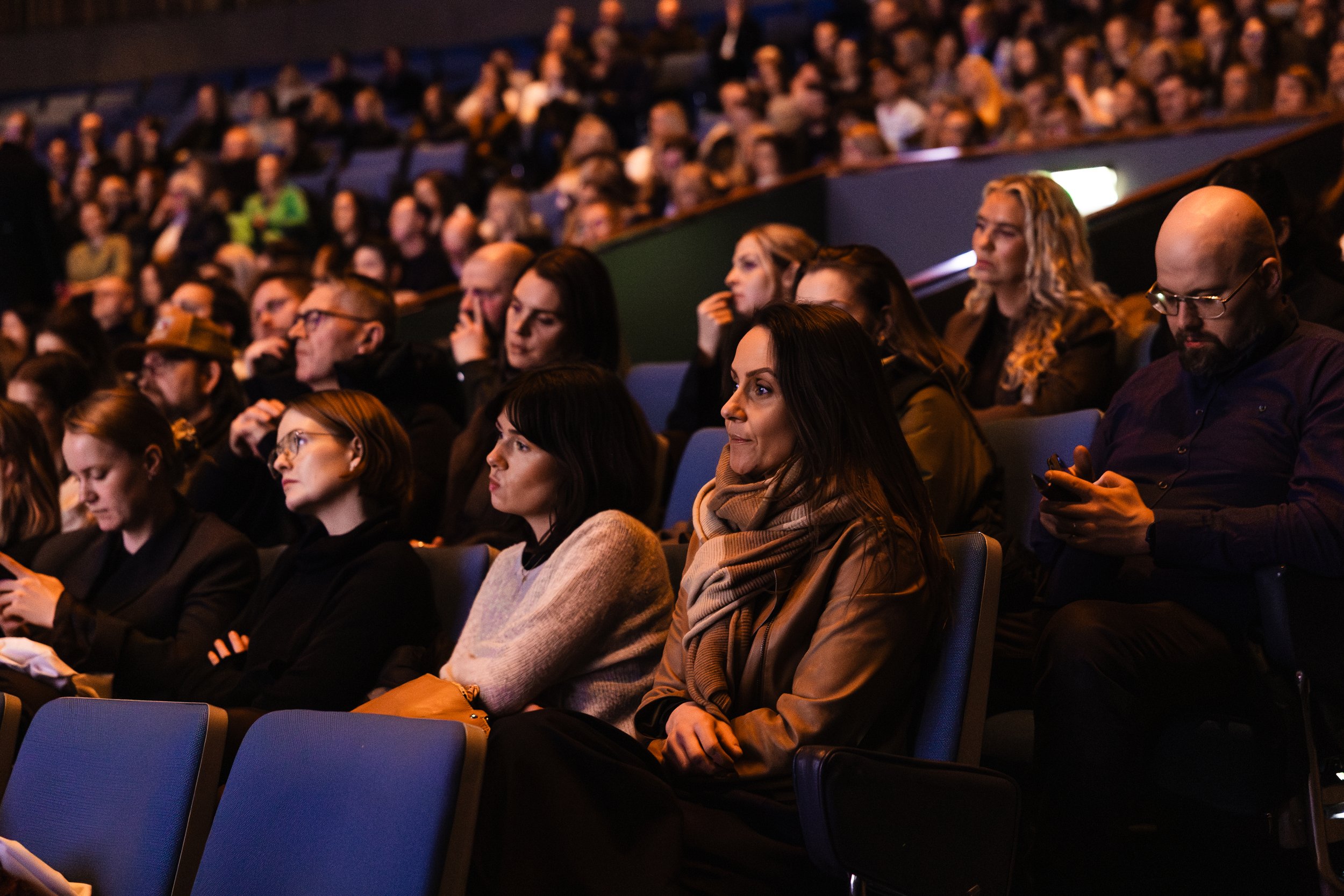 Audience seated in a theater listening to a presentation or performance, with some members using their phones.