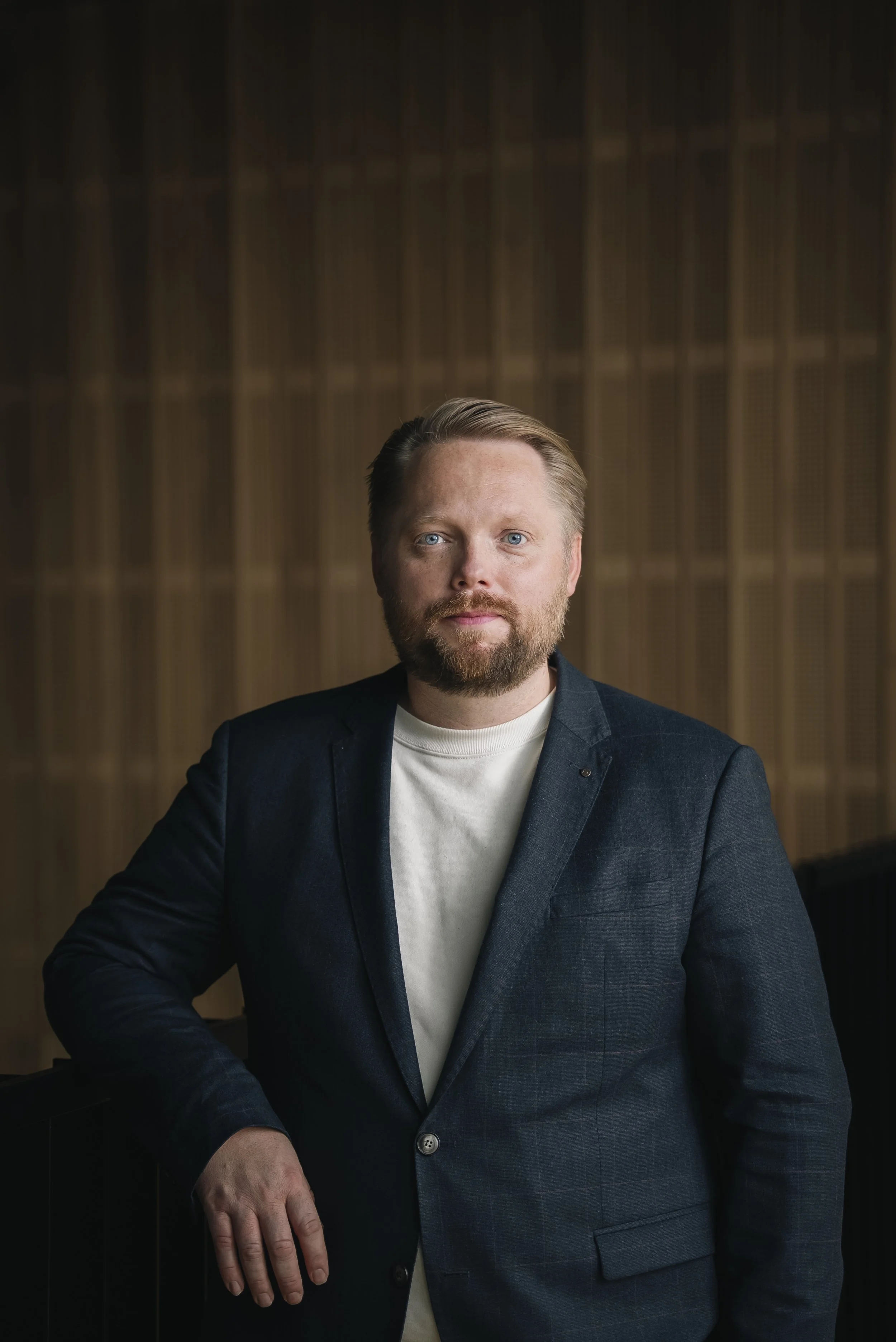 A man with a beard and blue eyes wearing a dark blue blazer and white shirt, posed with his hand resting on a dark surface, with a wooden wall in the background.