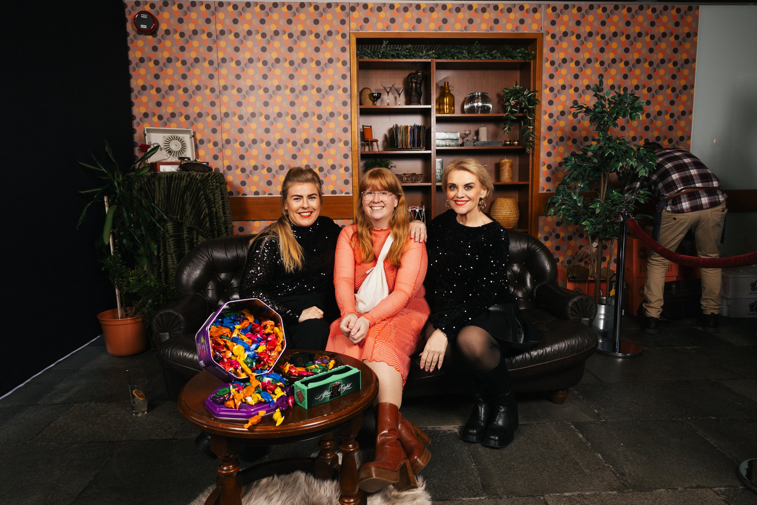 Three women sitting on a black leather couch in a living room, smiling at the camera. One woman has blonde hair and is wearing a black dress with sequins, the second has red hair and is wearing an orange dress, the third has blonde hair and is also i