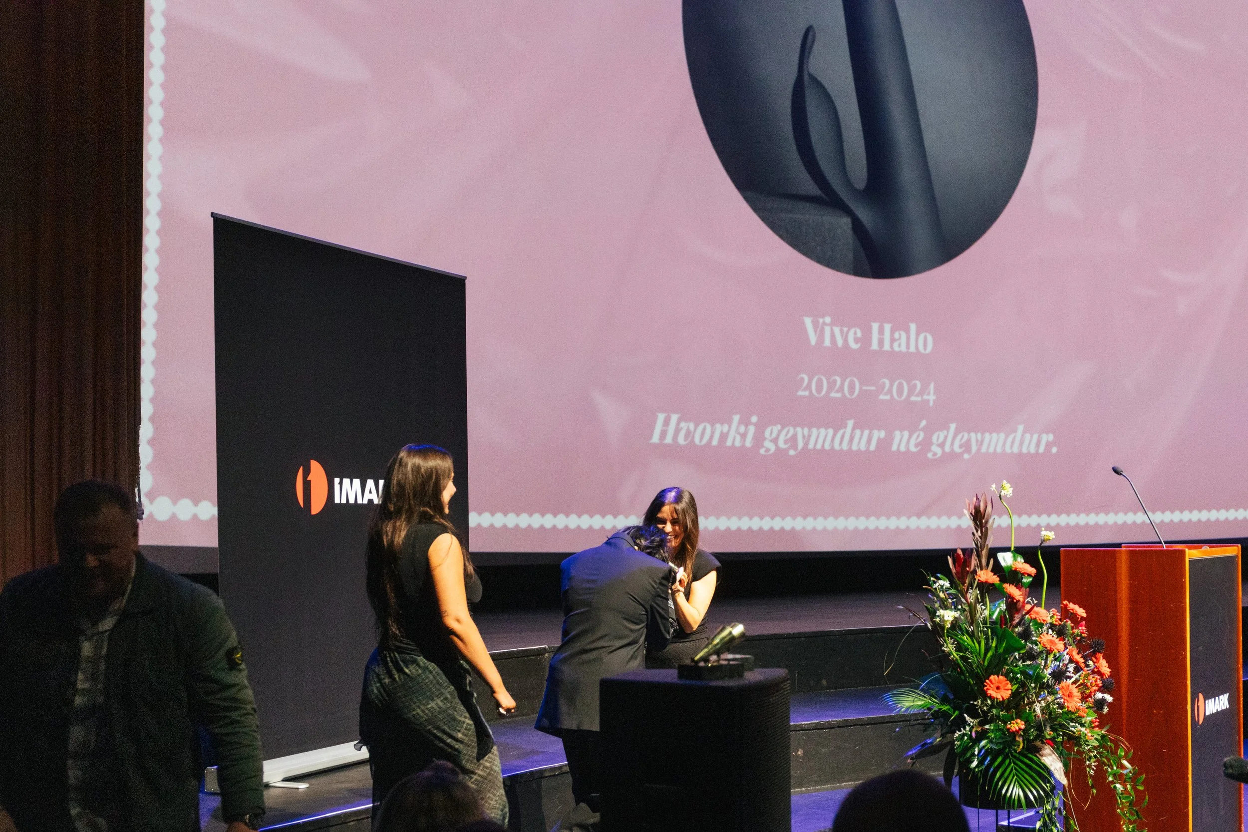 People on stage at an event with a large pink projection screen displaying text in a different language, decorated with flowers and plants.