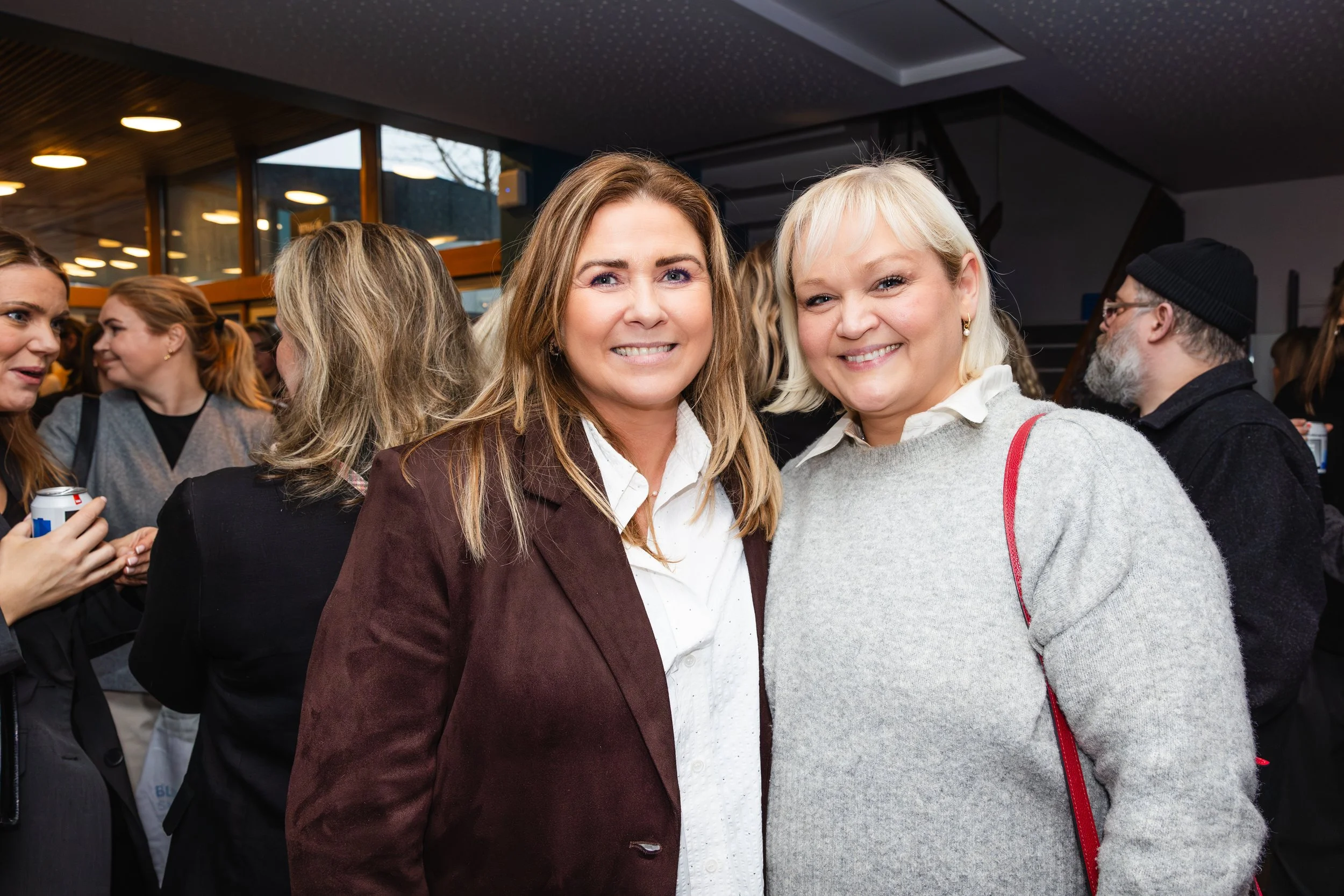 Two women smiling at a social gathering, standing close together. The woman on the left has shoulder-length brown hair and is wearing a white blouse with a dark blazer. The woman on the right has short blonde hair and is wearing a light-colored sweat