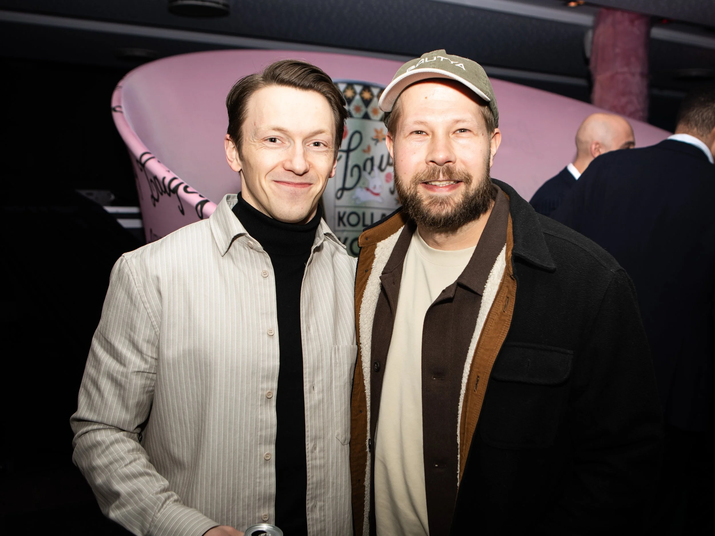 Two men are smiling for a photo, one wearing a striped shirt and the other in a jacket, with a pink airplane and a sign with the word 'Laut...'