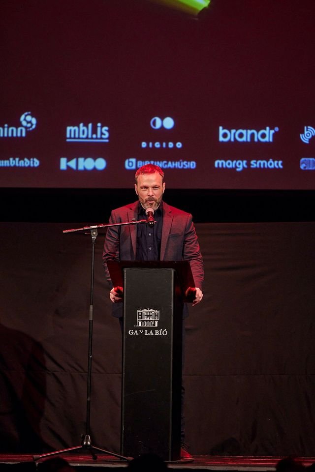 A man in a black suit stands behind a podium with a microphone, giving a speech at an event at GAV LA BIO. There is a large screen behind him displaying various logos.