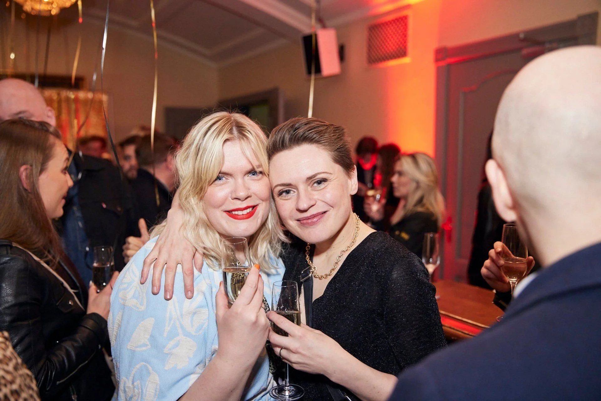 Two women smiling and posing with glasses of champagne at a lively party, surrounded by other guests in a warmly lit indoor setting.