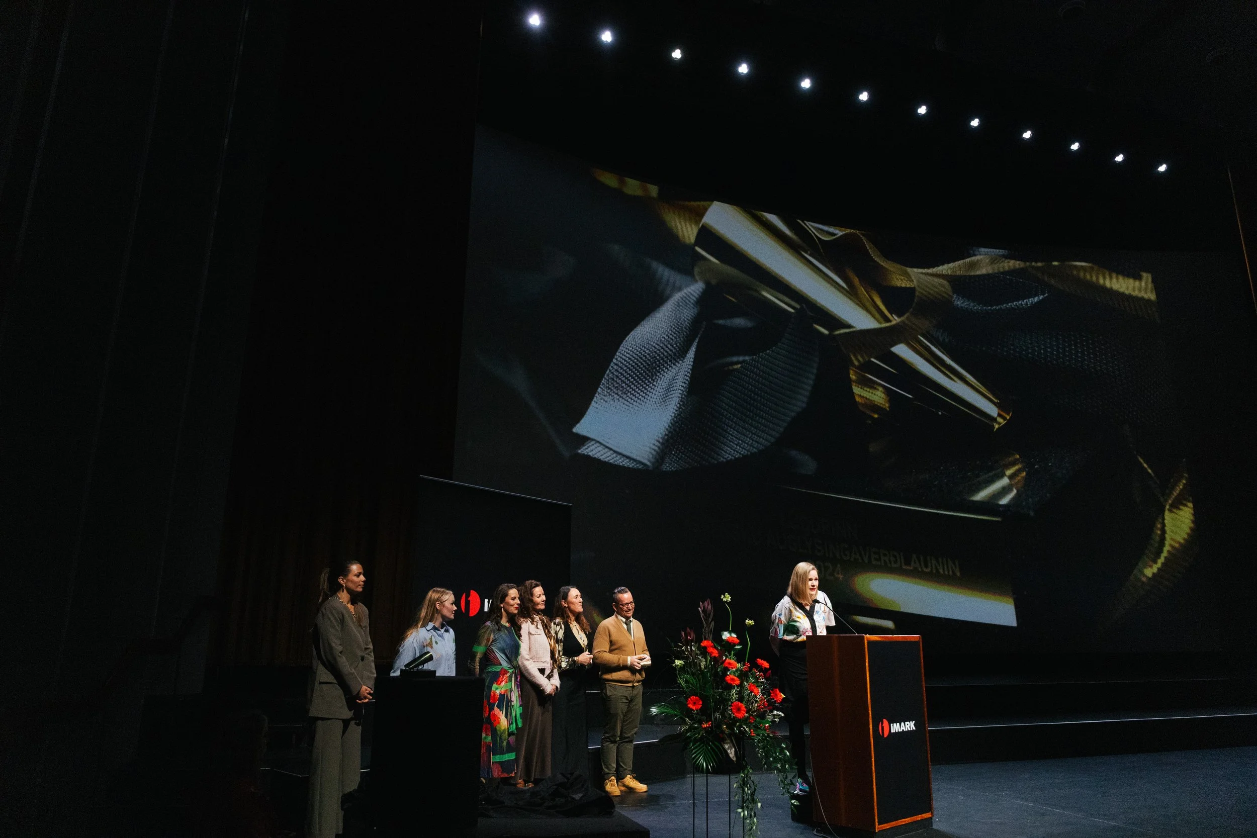A group of people standing on a stage in front of a large screen at a formal event or conference, with equipment and flowers arranged on the stage.