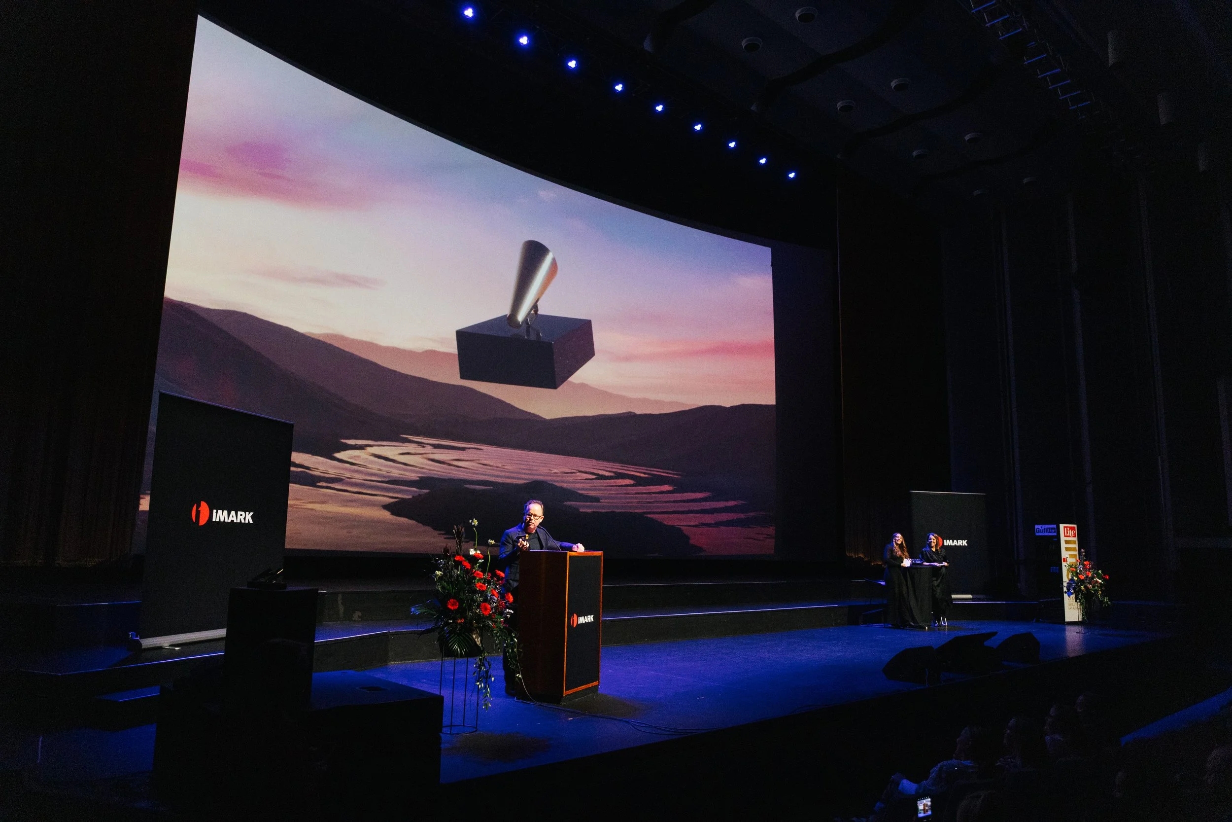 A stage with a large screen displaying a landscape with mountains and a river during sunset. Two people are standing at a podium with flowers, speaking to an audience. The stage has an IMARK logo, and two smaller screens on the stage display the IMAR