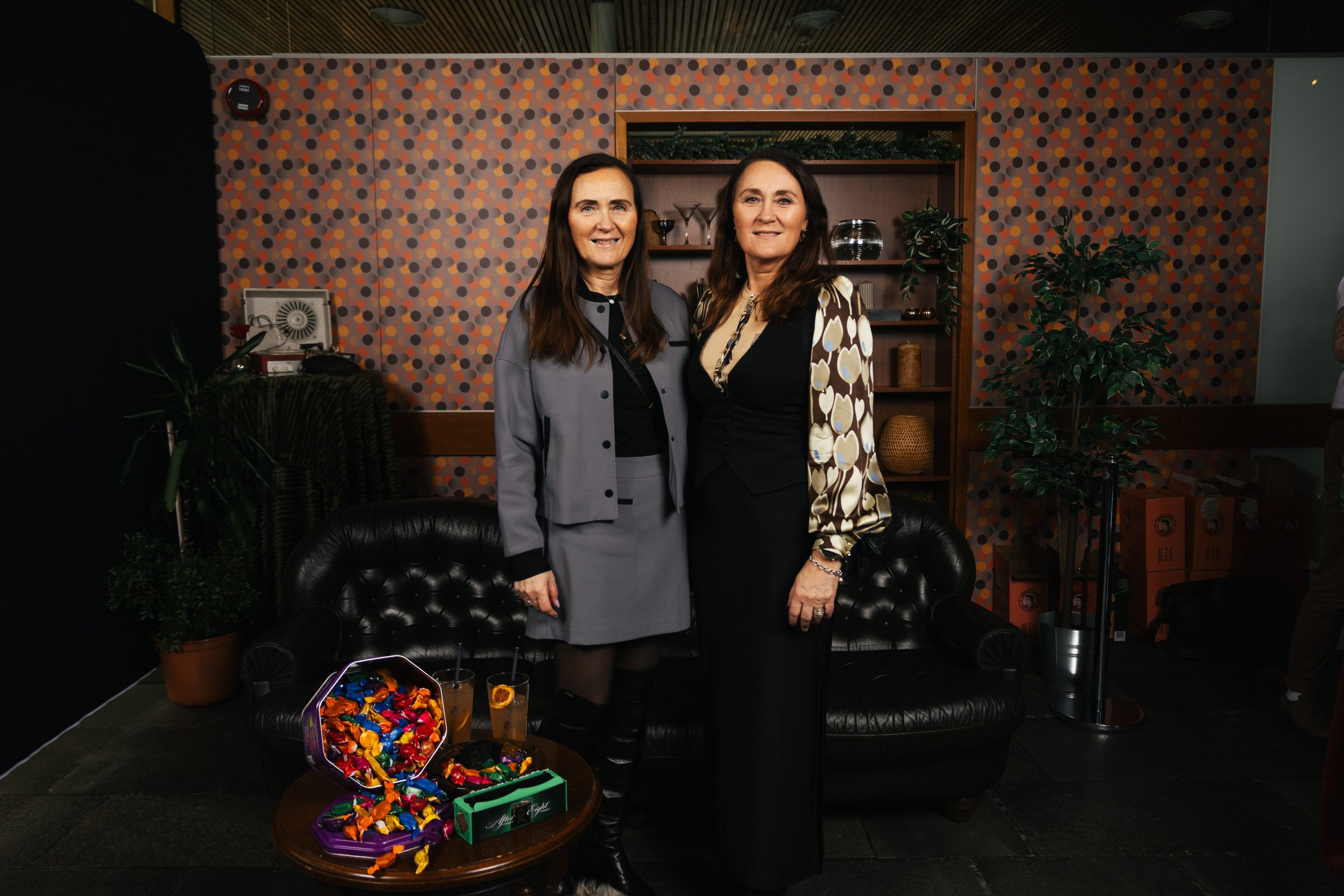 Two women standing together in an indoor setting. One woman is wearing a gray blazer and skirt, and the other is wearing a black top with a patterned jacket. They are smiling at the camera. Behind them is a patterned wallpaper, a wooden shelf with de