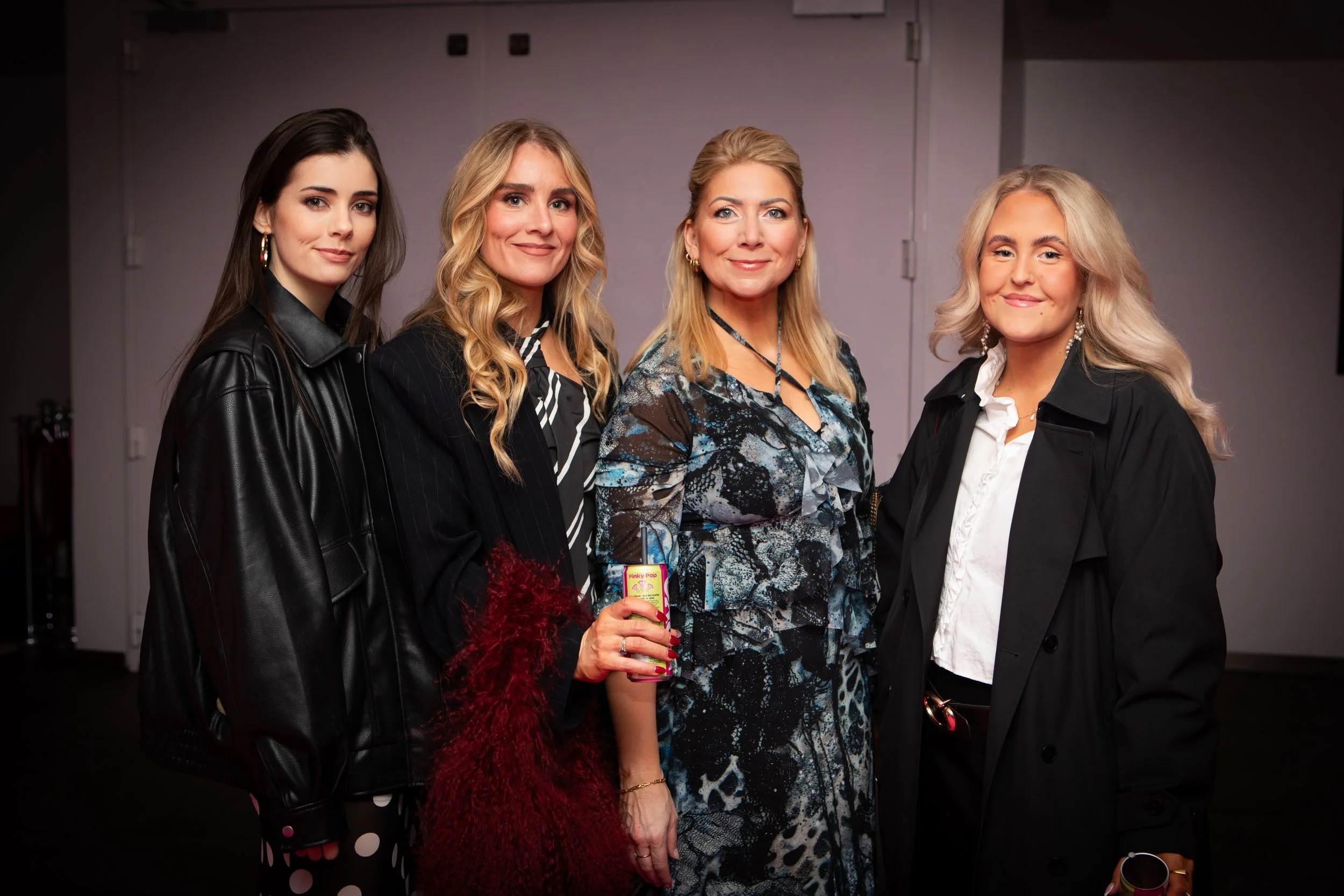 Four women standing together, smiling, in an indoor setting. The woman on the far left has long dark hair, wearing a black leather jacket. The second woman has blonde hair, wearing a dark blazer. The third woman has blonde hair styled up, wearing a p