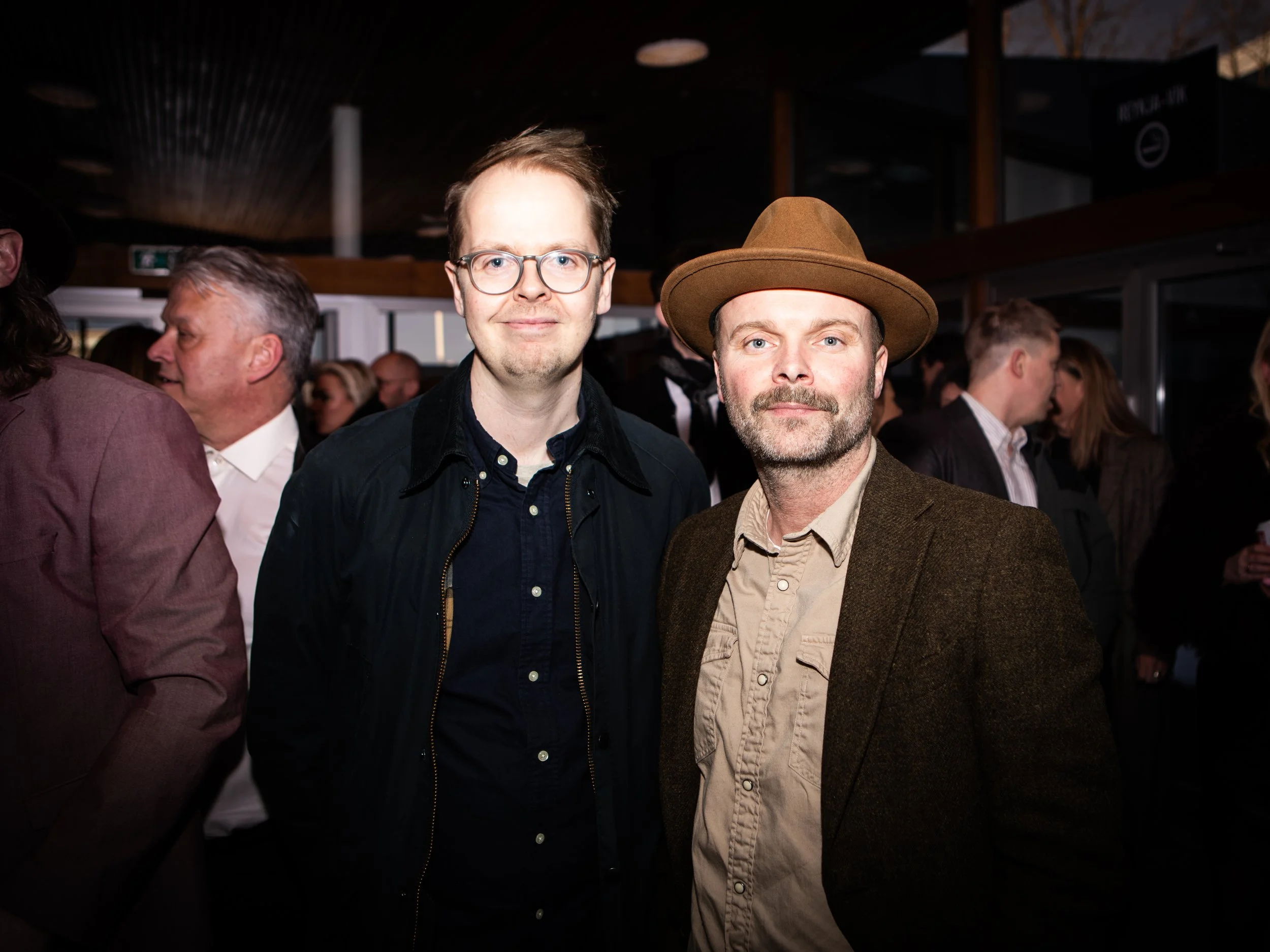 Two men standing close together at an indoor event, smiling, with a crowd of people in the background.
