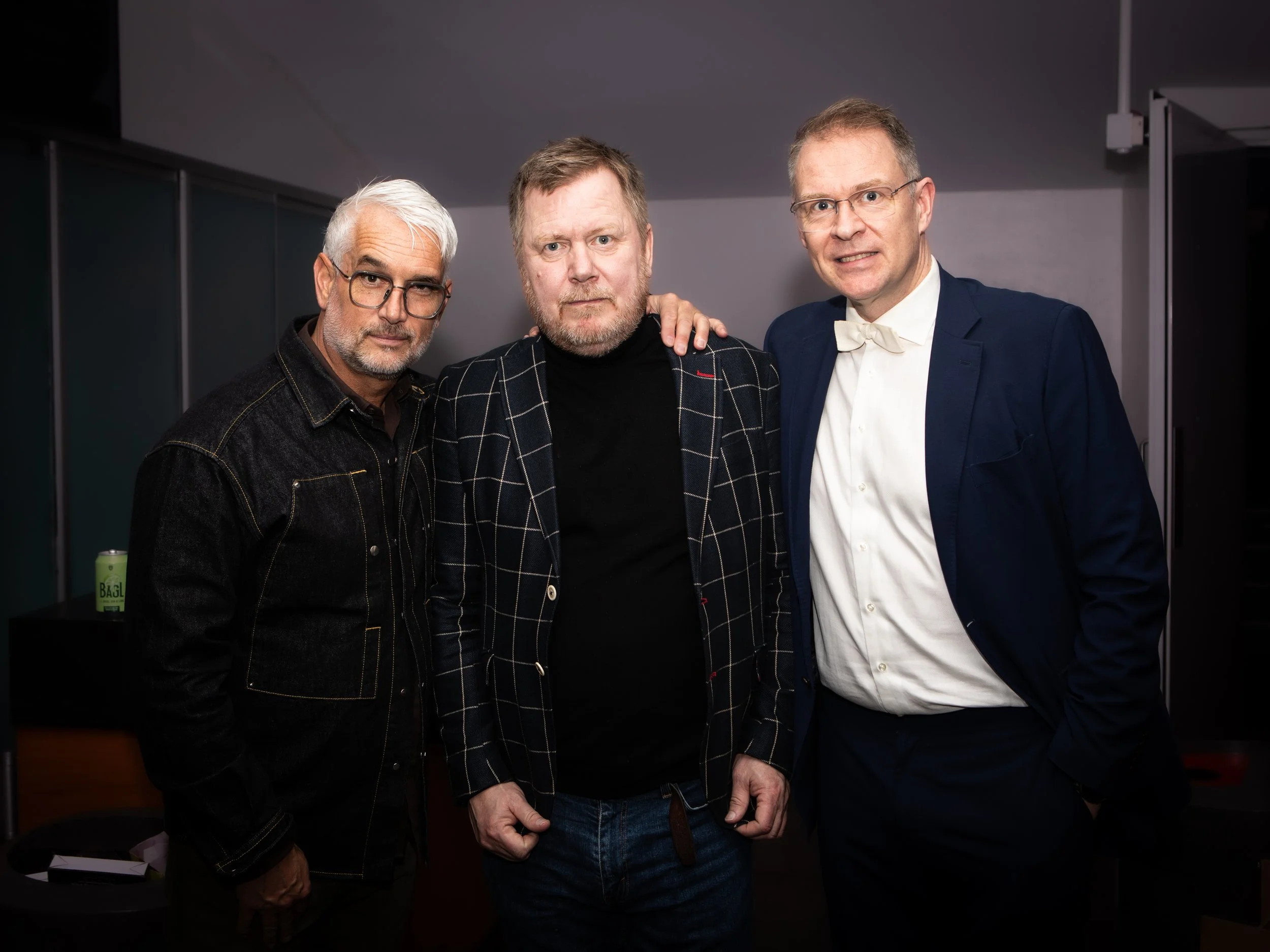 Three men standing together indoors, posing for a photo. The man on the left has white hair, glasses, and wears a dark denim jacket. The man in the middle has light brown hair and a beard, wearing a black shirt under a black and white checkered blaze