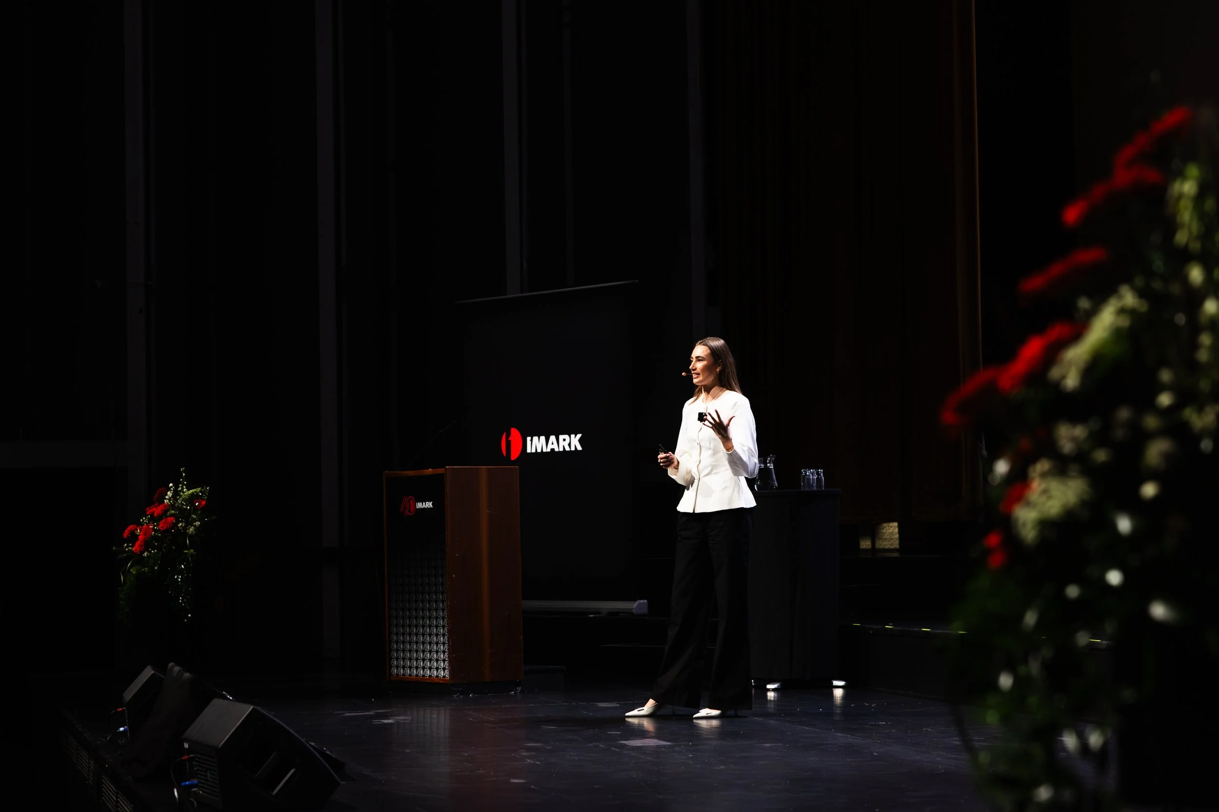 A woman with long dark hair, wearing a white blazer, black pants, and heels, standing on stage giving a presentation at an event, with a microphone headset, in front of a black screen displaying the 'IMARK' logo, decorated with red flowers.