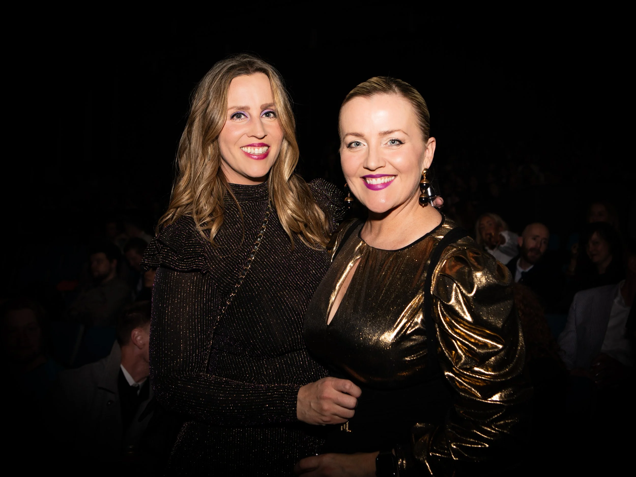 Two women smiling and dressed in glamorous black and gold outfits at a black background event, with one woman having long wavy hair and the other a short hairstyle.