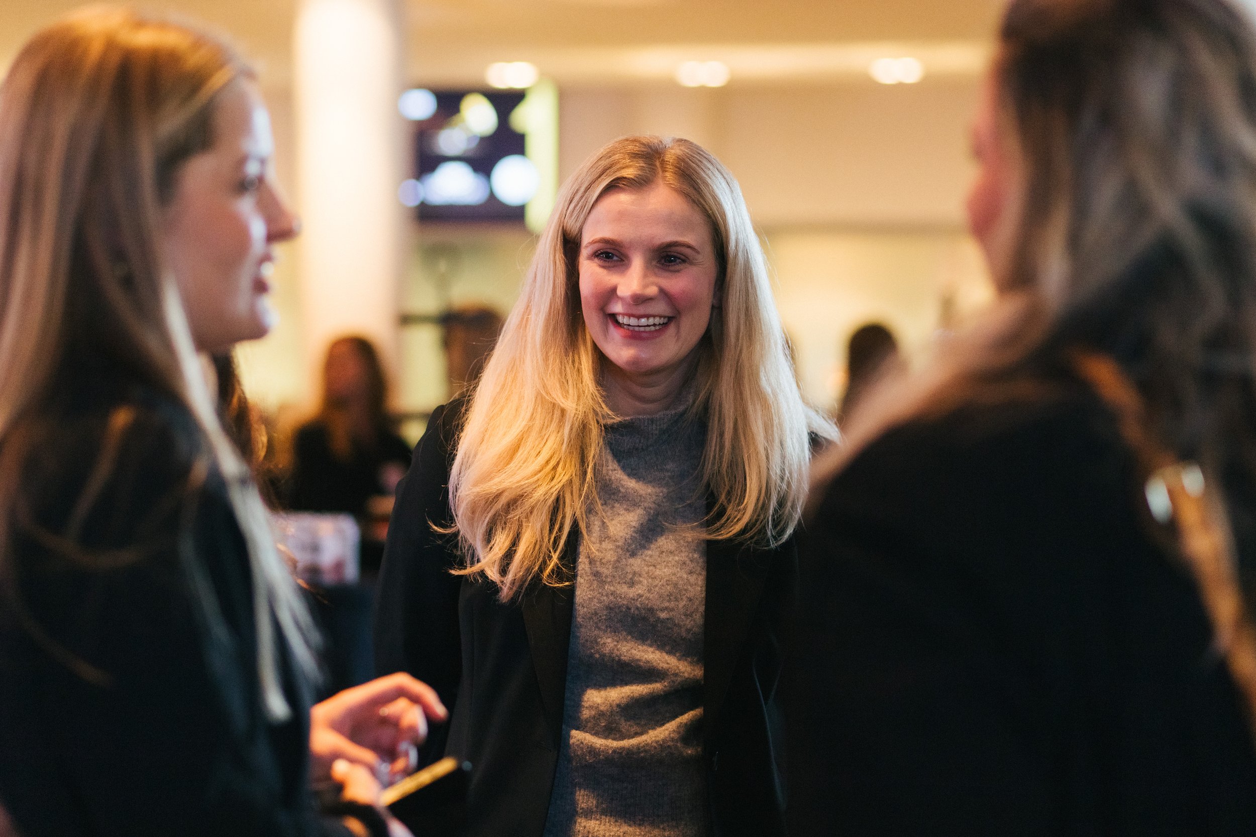 Three women having a conversation in a well-lit indoor setting, with the central woman smiling.