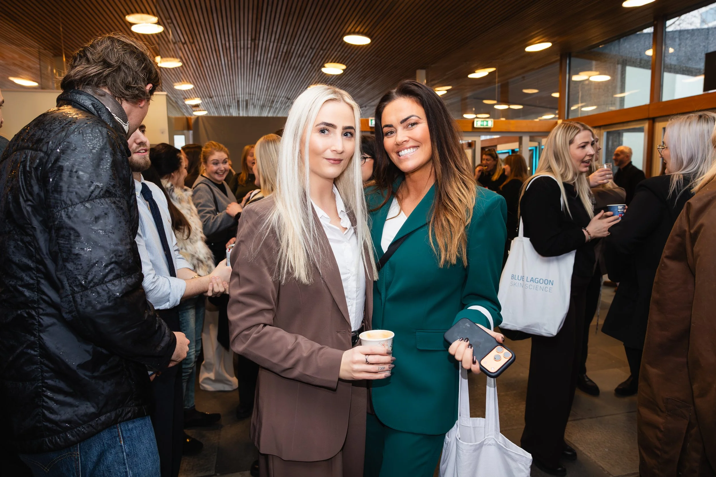 Two women smiling and standing close together in a crowded indoor event, one holding a coffee cup and a smartphone, with people talking and mingling in the background.