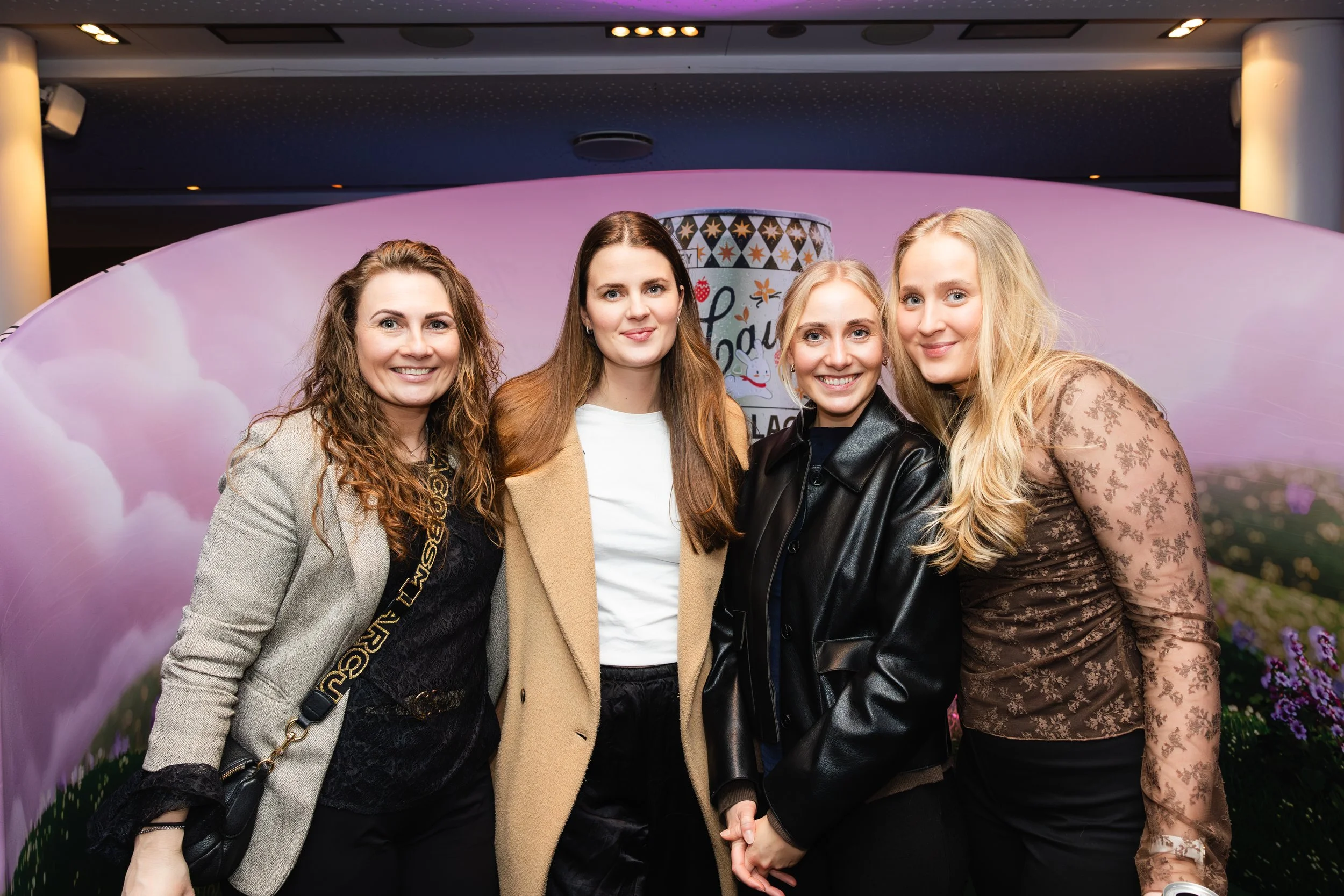 Four women standing close together indoors, smiling, with a pink floral backdrop behind them.