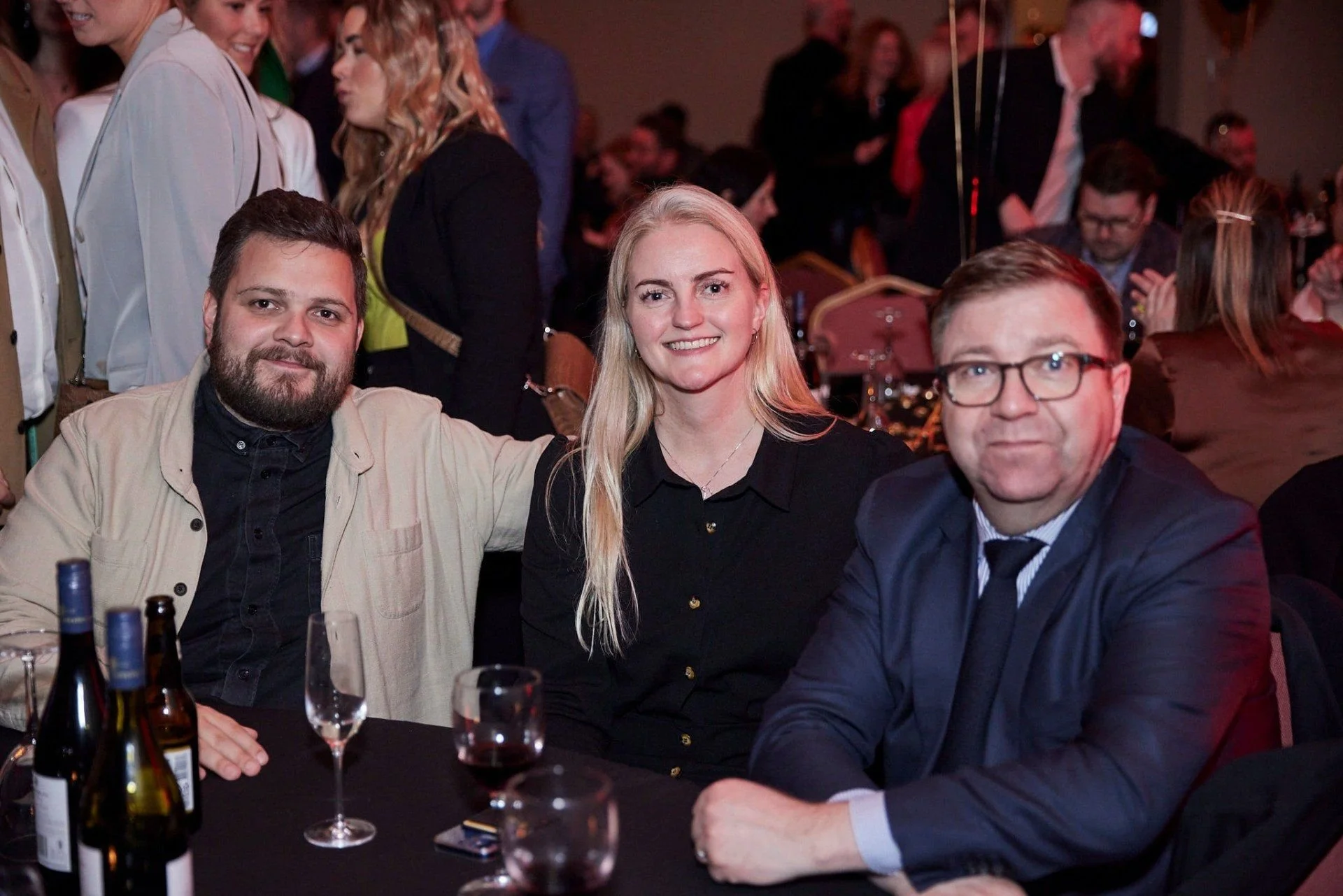 Three people sit at a table in a crowded event, smiling at the camera. There are bottles and glasses on the table, and other people are visible in the background.