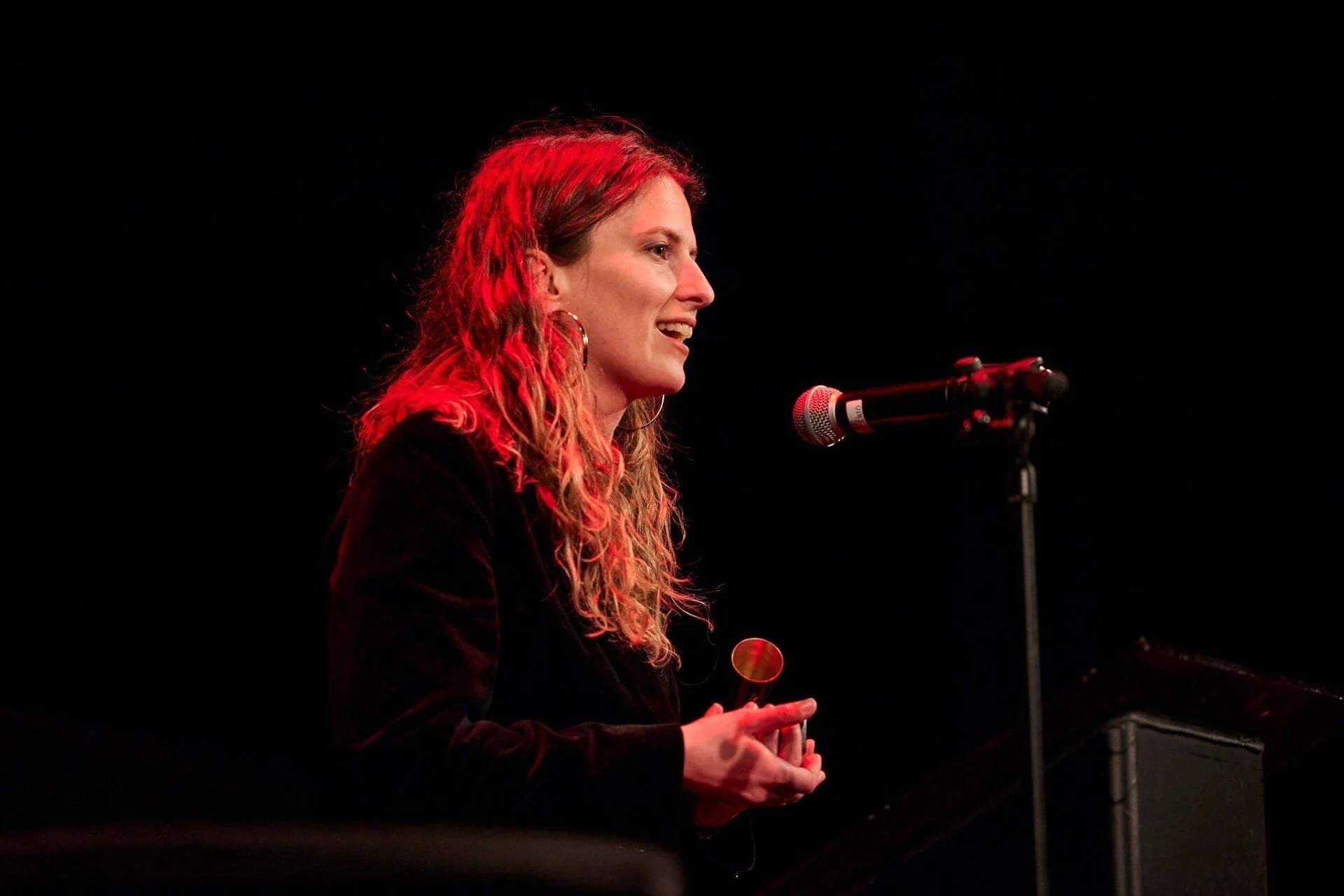 A woman with red curly hair speaking into a microphone on stage, with a dark background and a piano nearby.