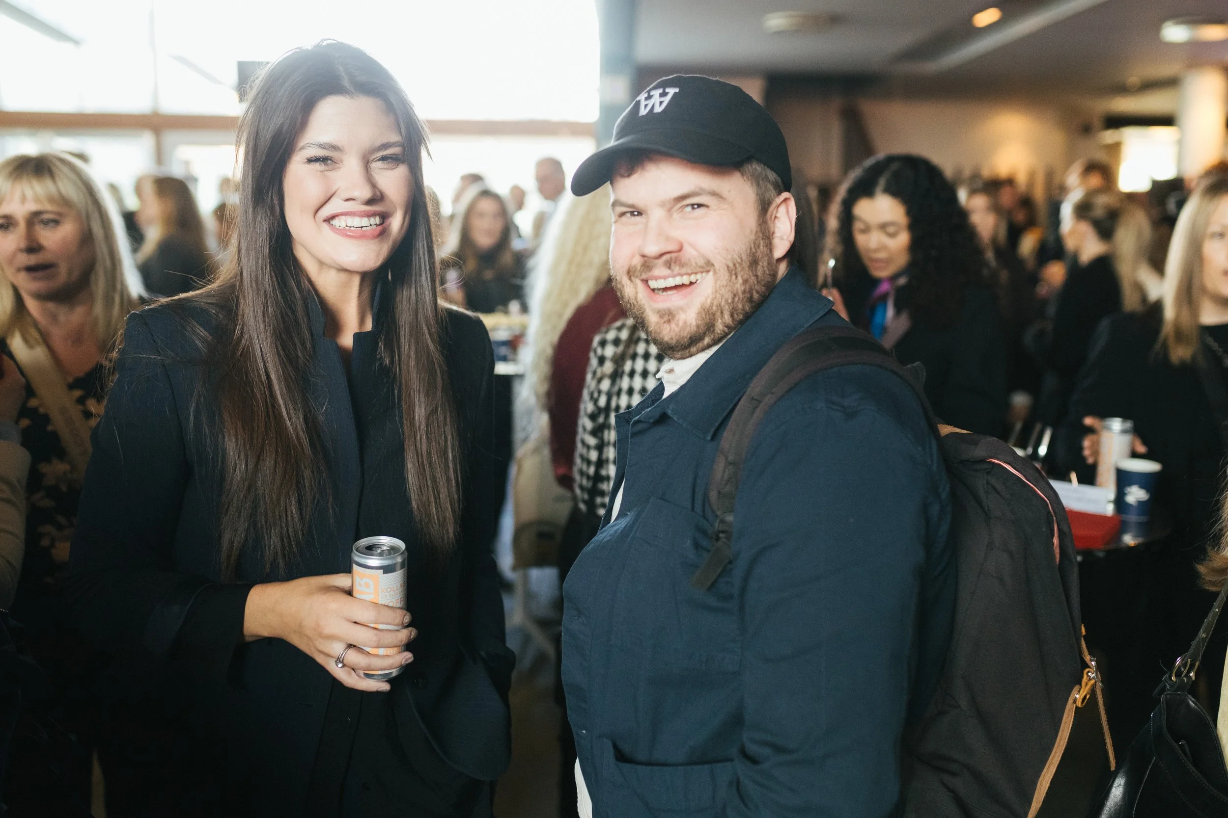 A woman and a man smiling at an indoor social event, with many people in the background.