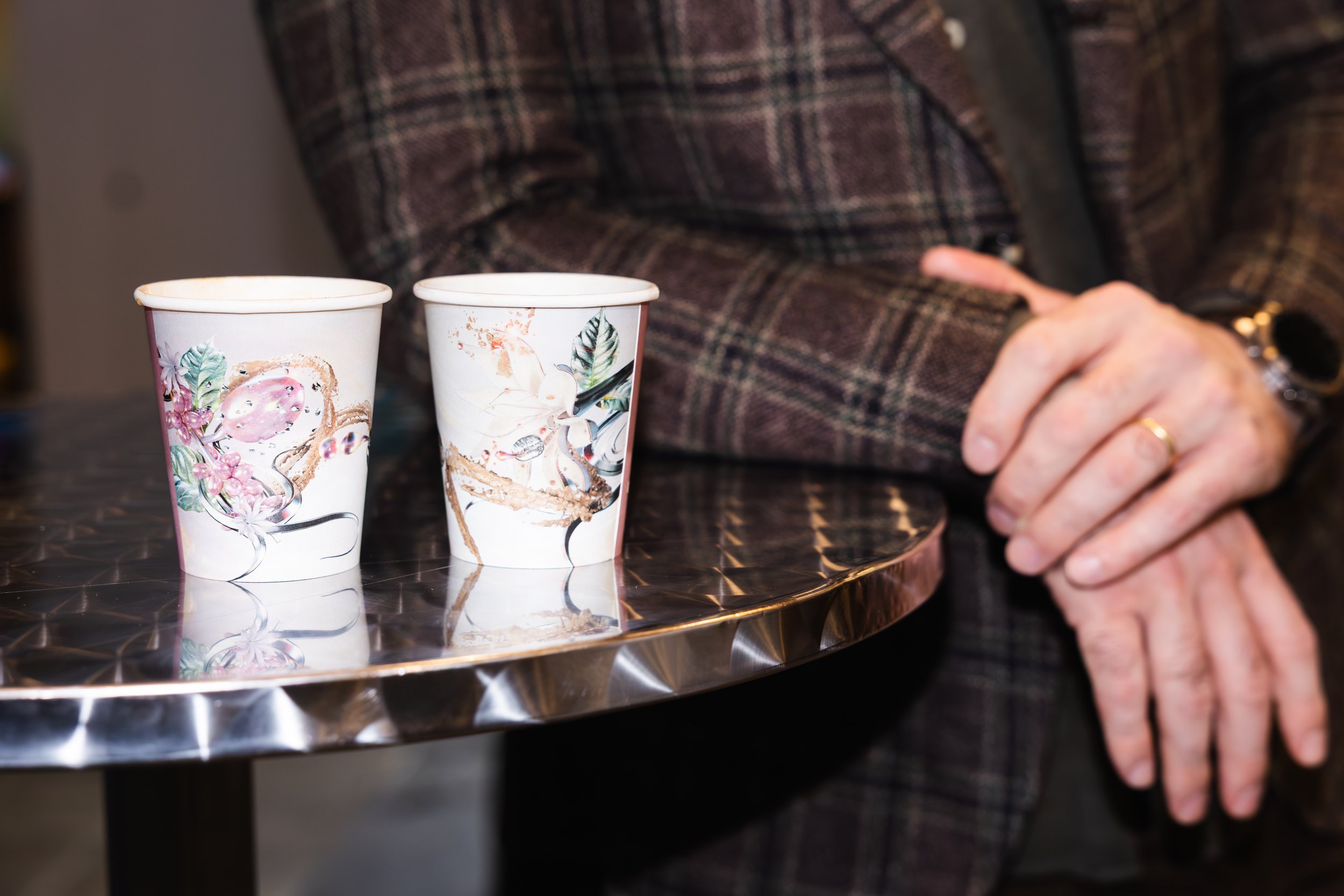 Two disposable coffee cups with floral and animal designs are placed on a reflective surface. In the background, a person's hand with rings and a gold chain watch is resting on the table, wearing a plaid jacket.