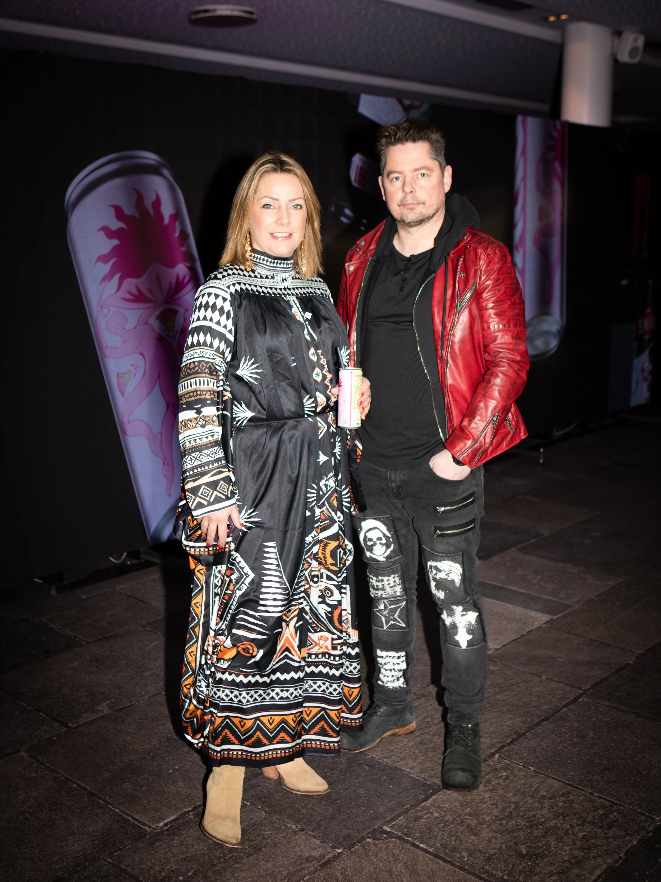 A woman and a man standing together in front of a dark backdrop, with pink and purple graphics on the left side of the image. The woman is wearing a colorful, patterned dress with beige boots, and the man is wearing a red leather jacket, black shirt,