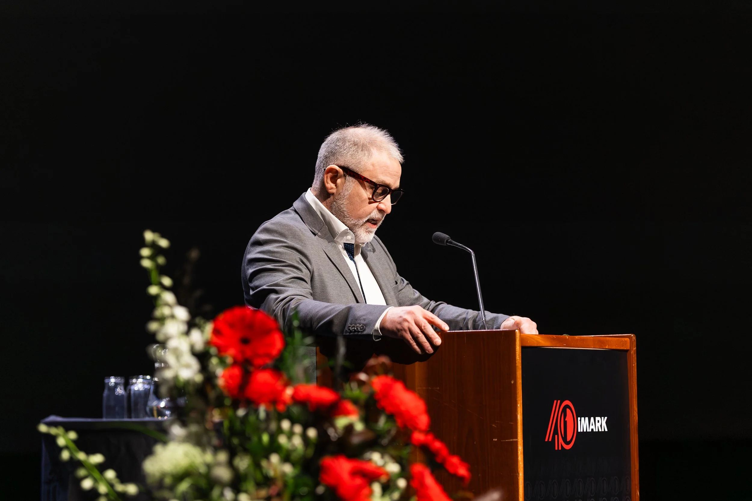 A man with gray hair and glasses, wearing a gray suit, is speaking at a podium during an event. There are red and white flowers in the foreground and a black background.