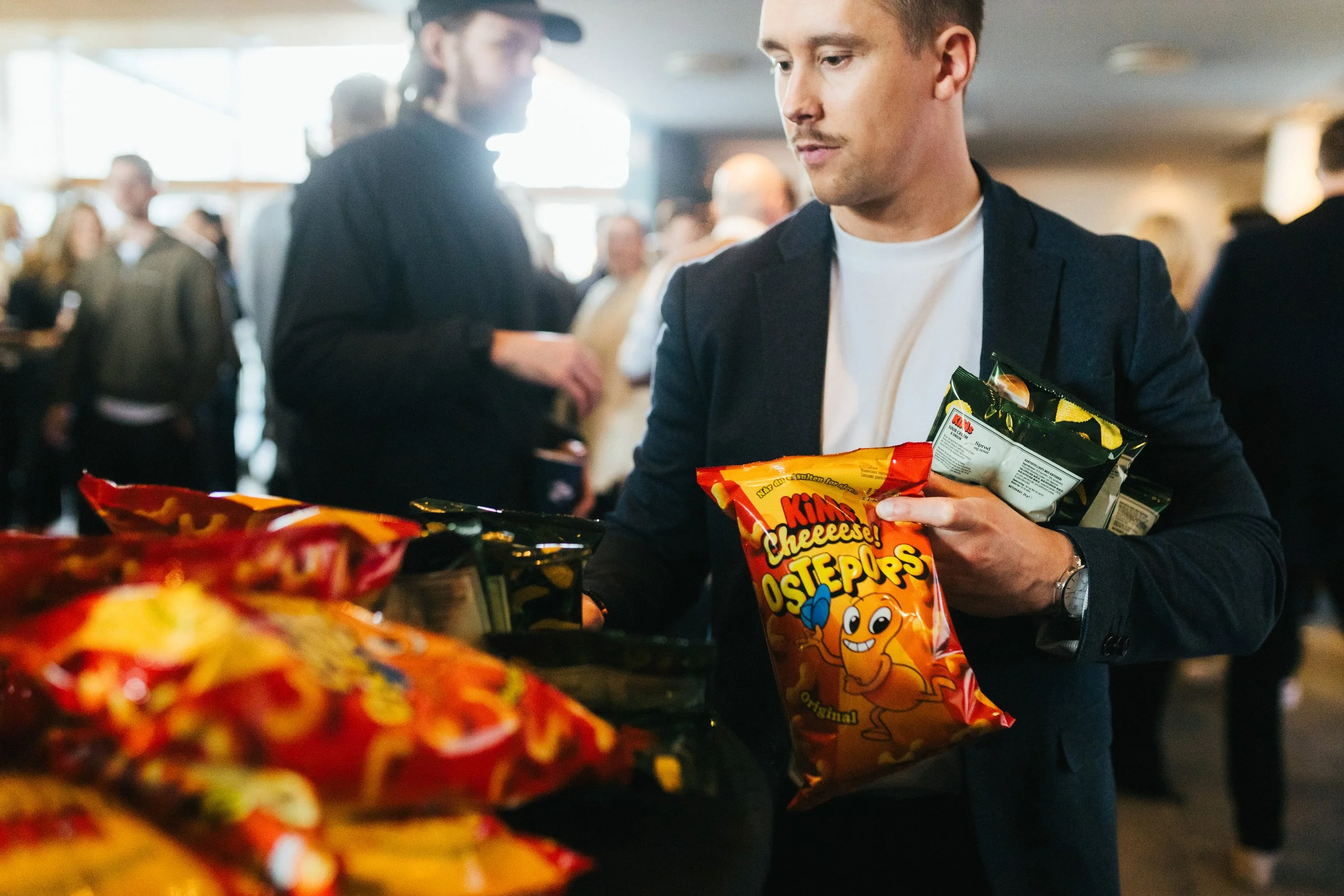 A young man in a suit and white shirt shopping for snack foods at a store, holding a bag of Cheeester Os in his hand.