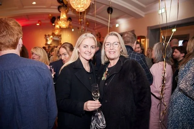 Two women smiling at a social gathering in an elegant room, one holding a glass of champagne, surrounded by other guests.