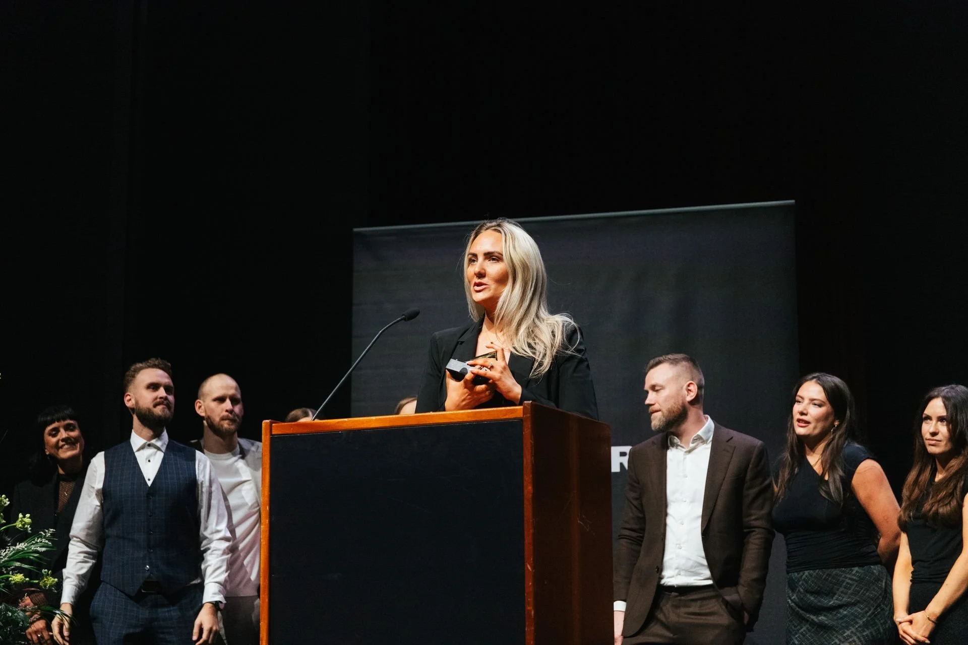 Woman speaking at podium surrounded by people at a formal event in a dark auditorium.