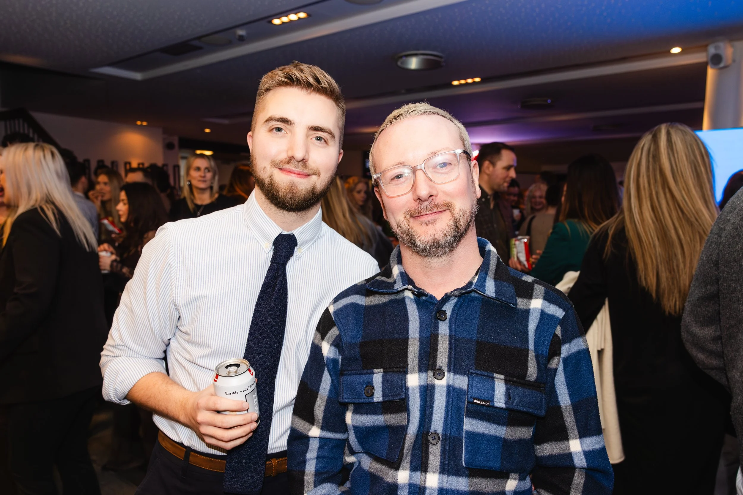 Two men posing together at a social event. The man on the left has short, light brown hair, a beard, and is wearing a white shirt with a dark tie, holding a soda can. The man on the right has short, light hair, glasses, and a beard, wearing a blue an