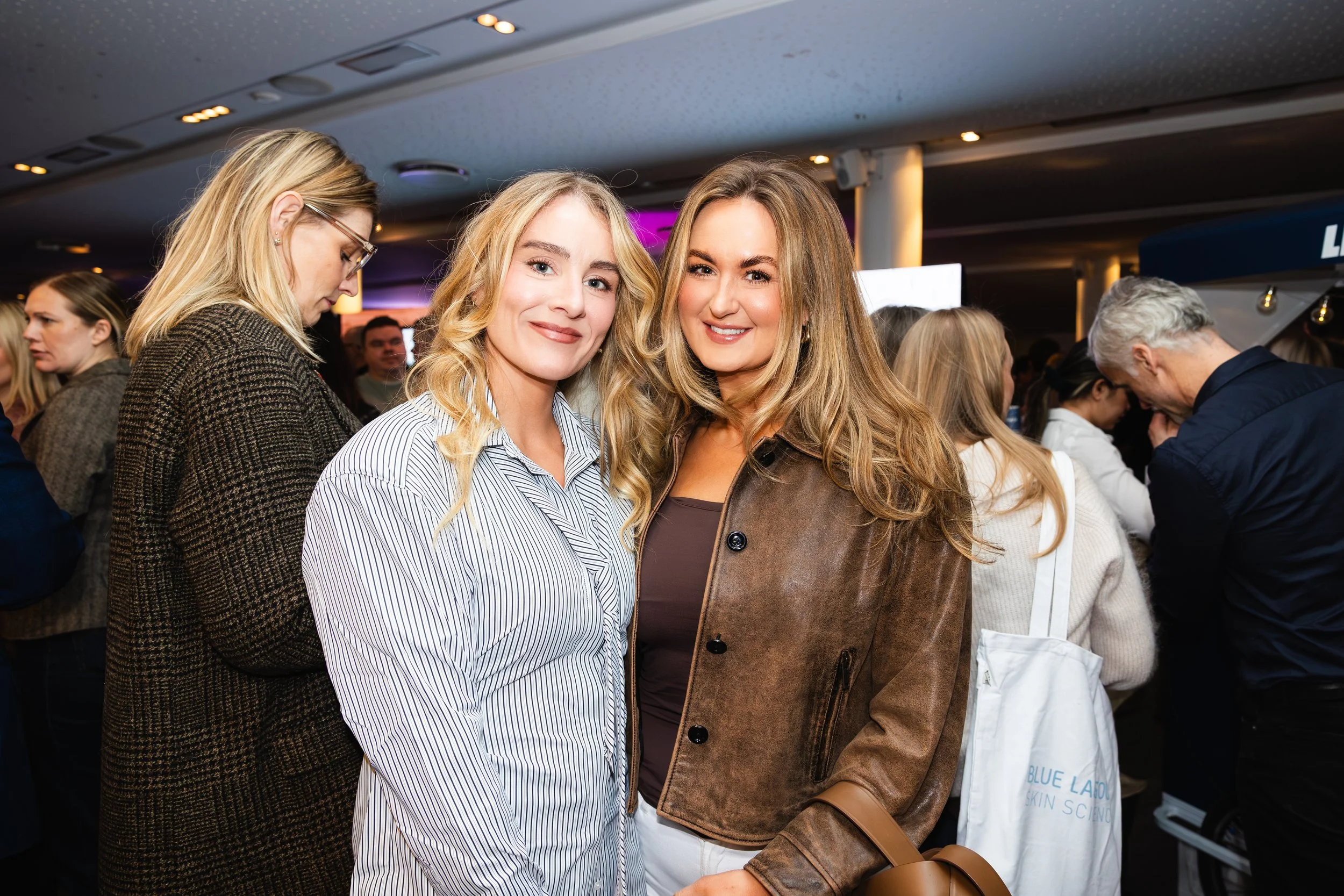 Two women smiling in a crowded indoor setting, possibly at a social event.