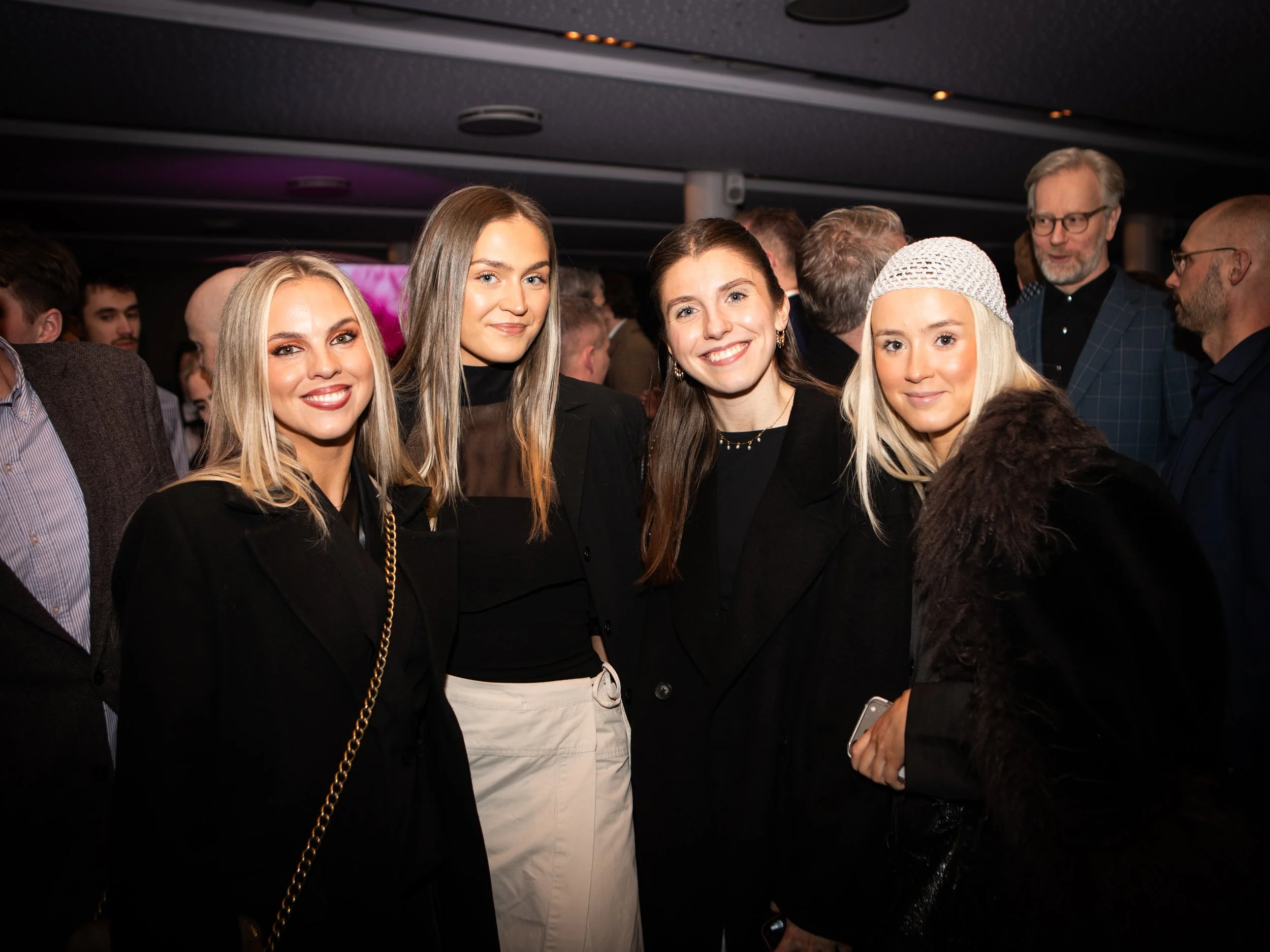 Four young women smiling at a social event, standing close together in a crowded indoor setting.
