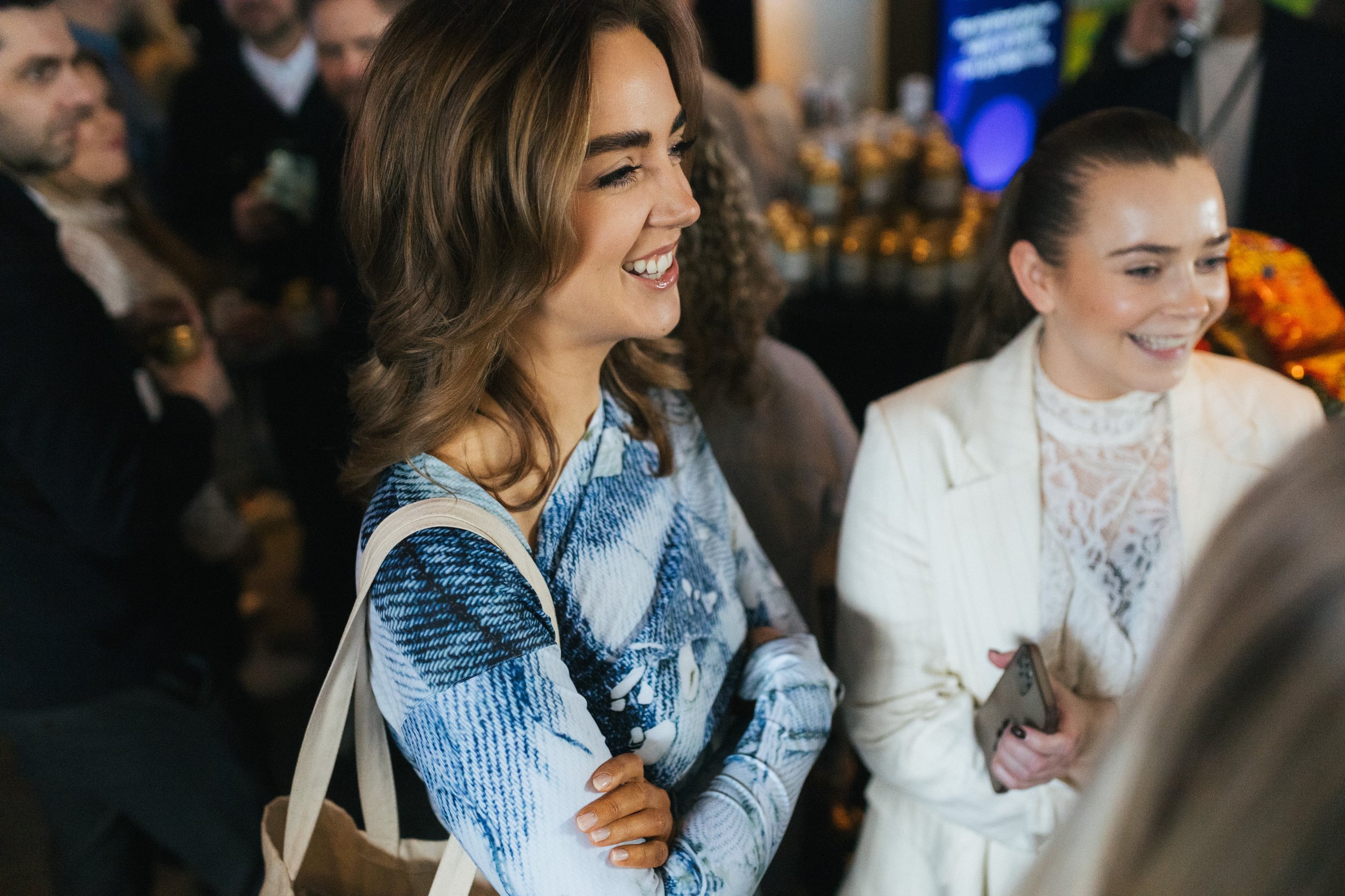 Two women smiling and sitting together at an indoor event with other people in the background.