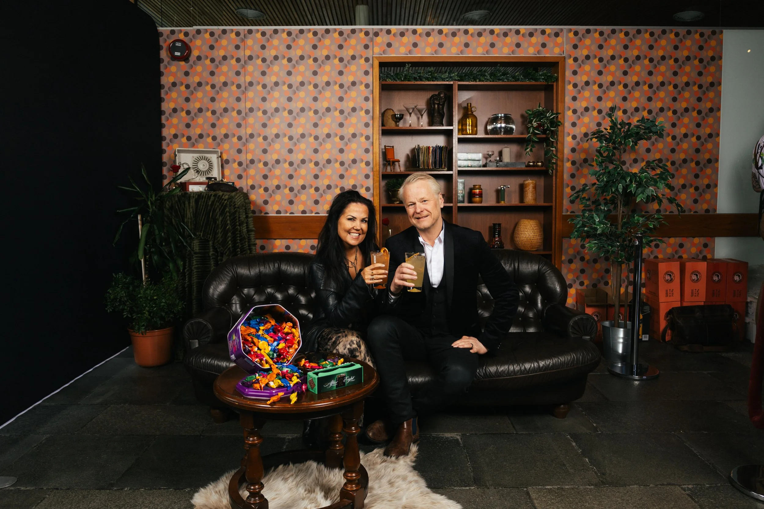 A smiling woman and man sitting on a black leather sofa indoors, holding drinks. The woman is wearing a black outfit and patterned tights, and the man is in a dark suit. In front of them is a coffee table with a purple box of candies. The background 