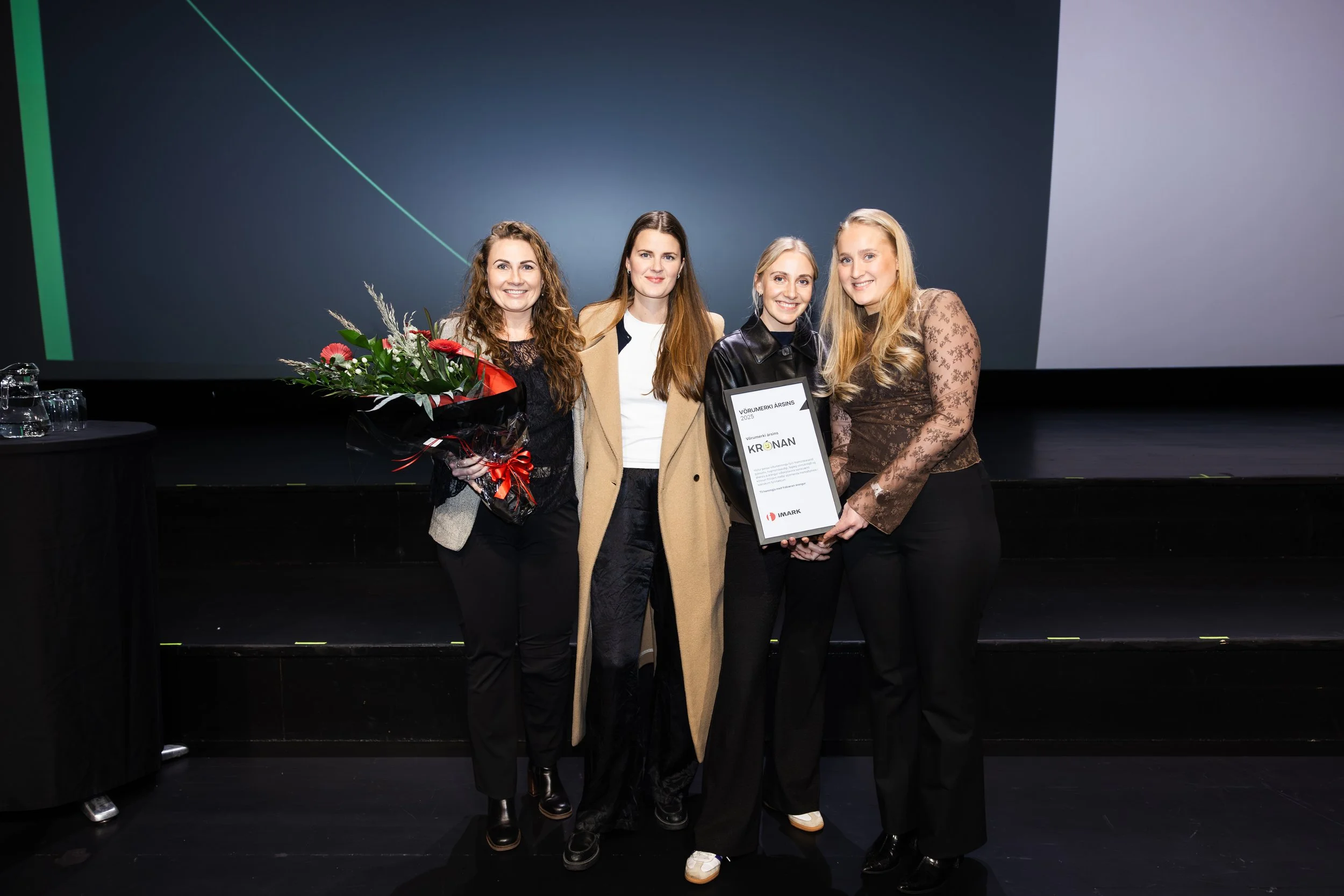 Four women standing together on a stage, smiling, with one holding a framed certificate and a bouquet of flowers.