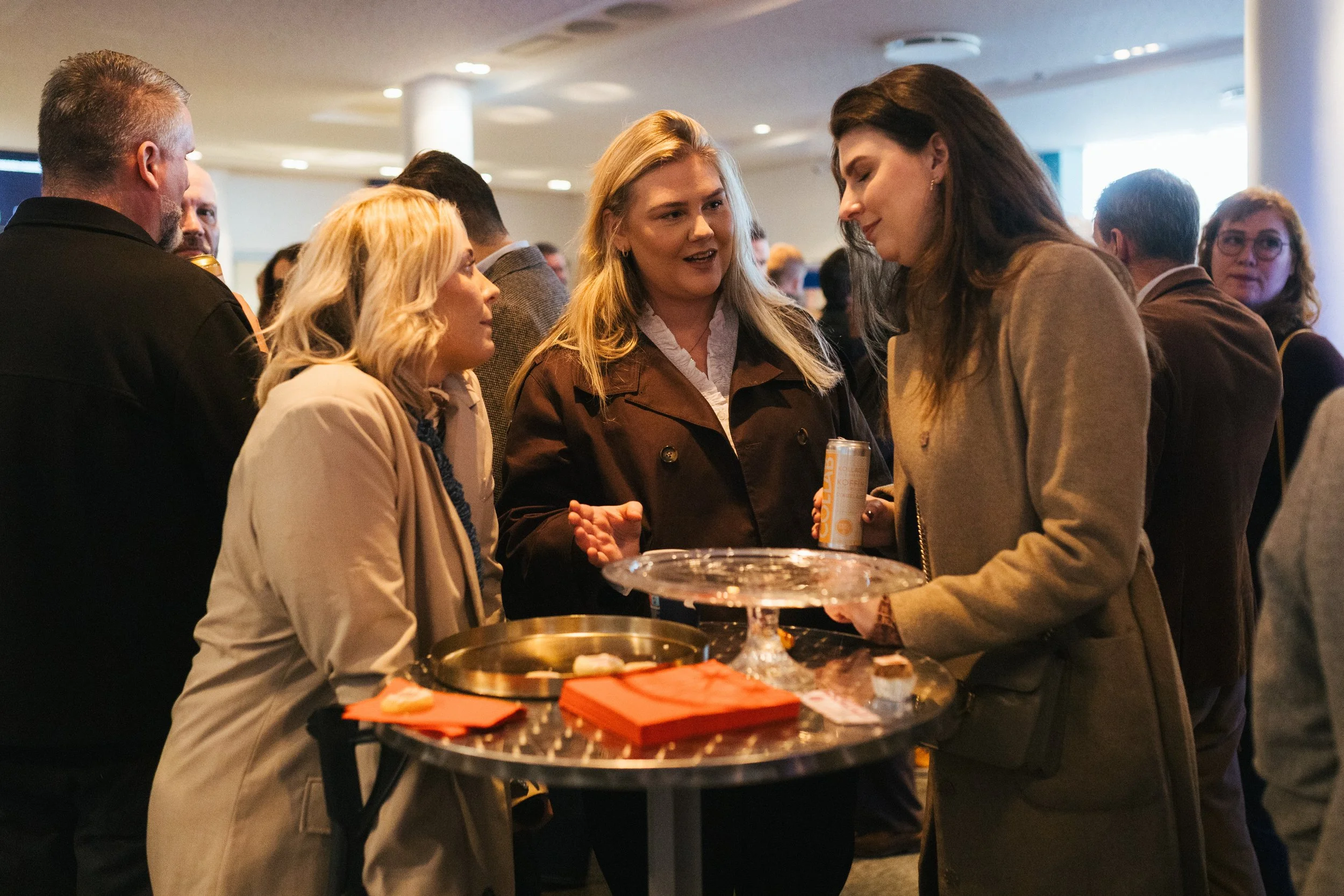 Group of diverse adults engaged in conversation at an indoor gathering or event, standing around a round table with snacks and drinks.