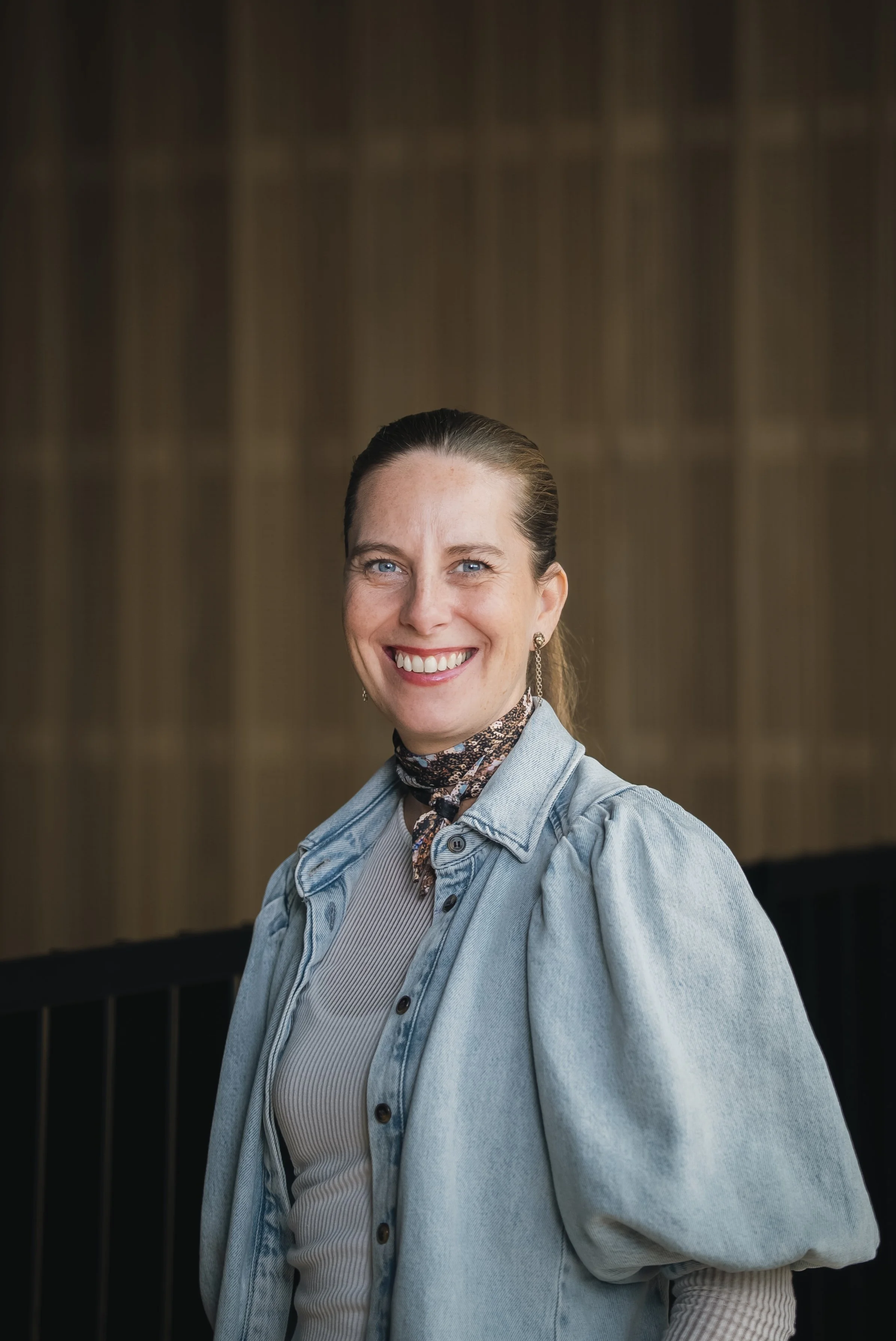 A smiling woman with blue eyes and dark hair pulled back, wearing a denim jacket over a beige ribbed top, standing in front of a dark and blurred background.