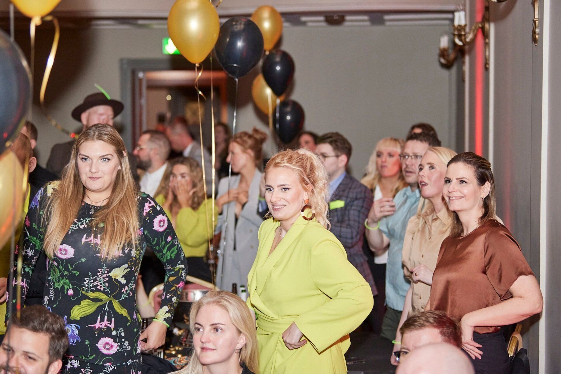 People gathered at an indoor event with balloons, some are standing and some seated, engaged in conversation and watching the event.