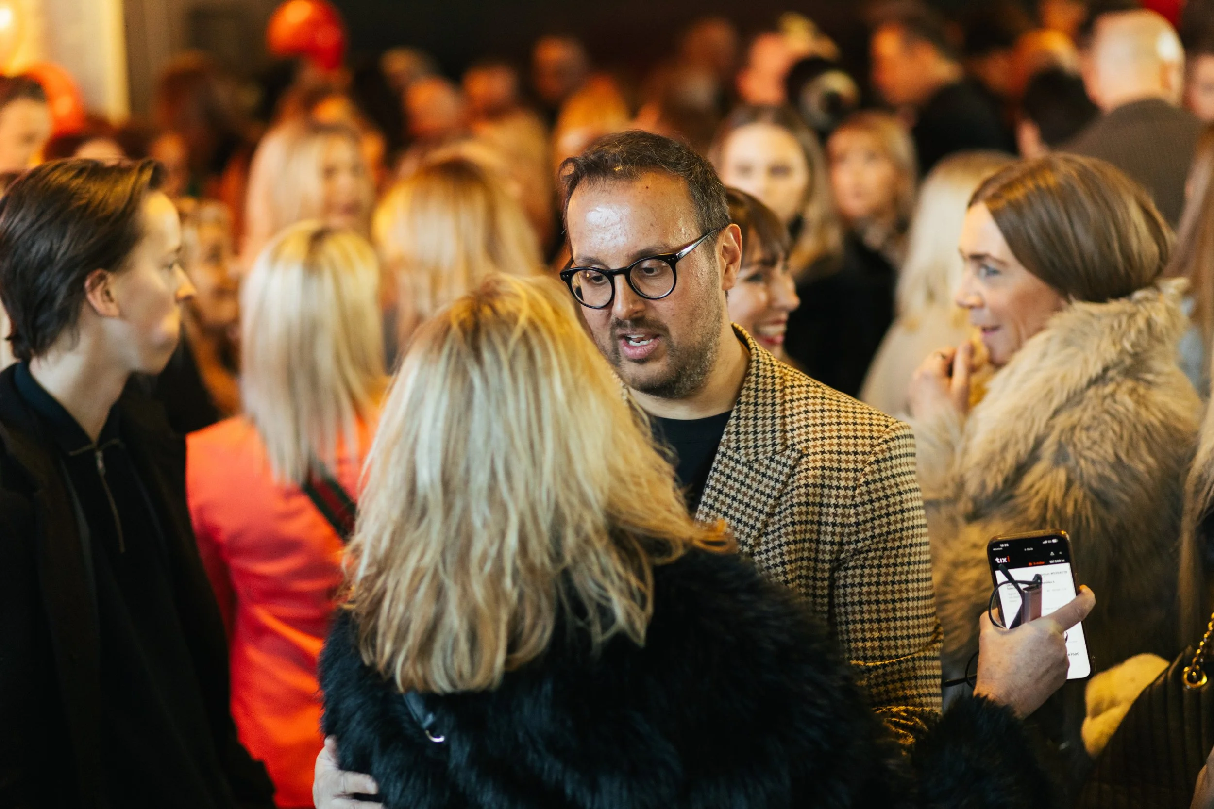 Group of people gathered at an indoor event, engaging in conversation, with a man in glasses and a patterned blazer in focus in the center.