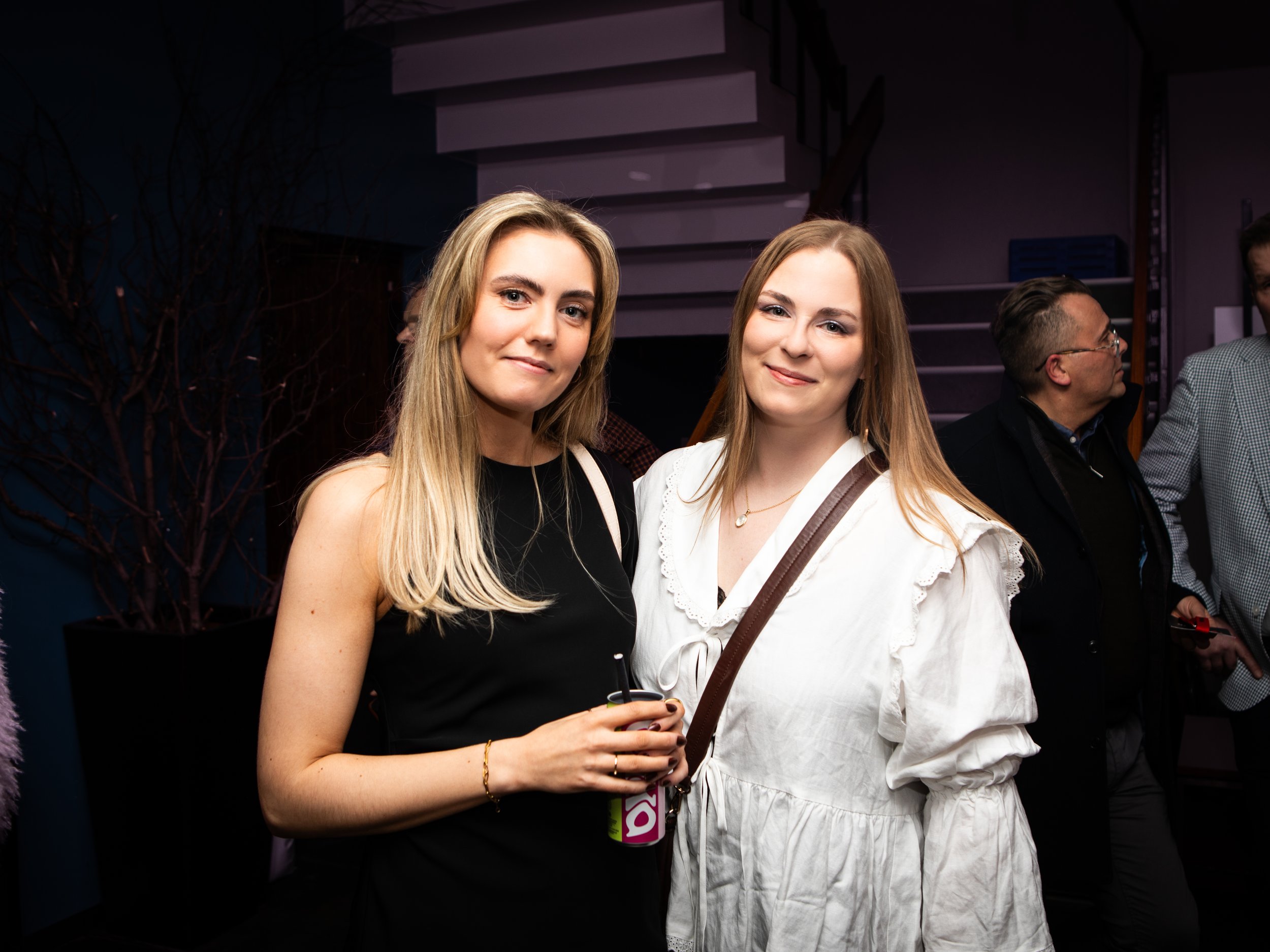 Two young women with long hair smiling at the camera at an indoor event, one wearing a black top and the other in a white dress. A man with glasses is partially visible in the background, along with a dark background and stairs.