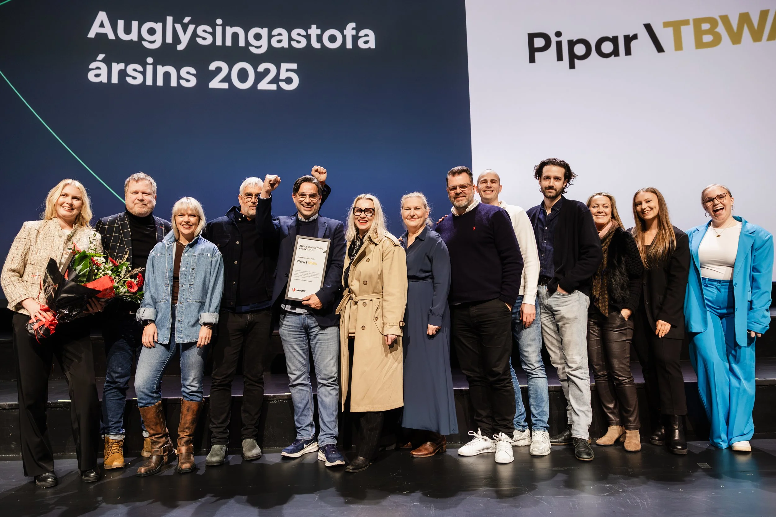 A group of people on stage celebrating, with some holding flowers and a framed certificate. Behind them is a large screen with text in a Scandinavian language and the year 2025.