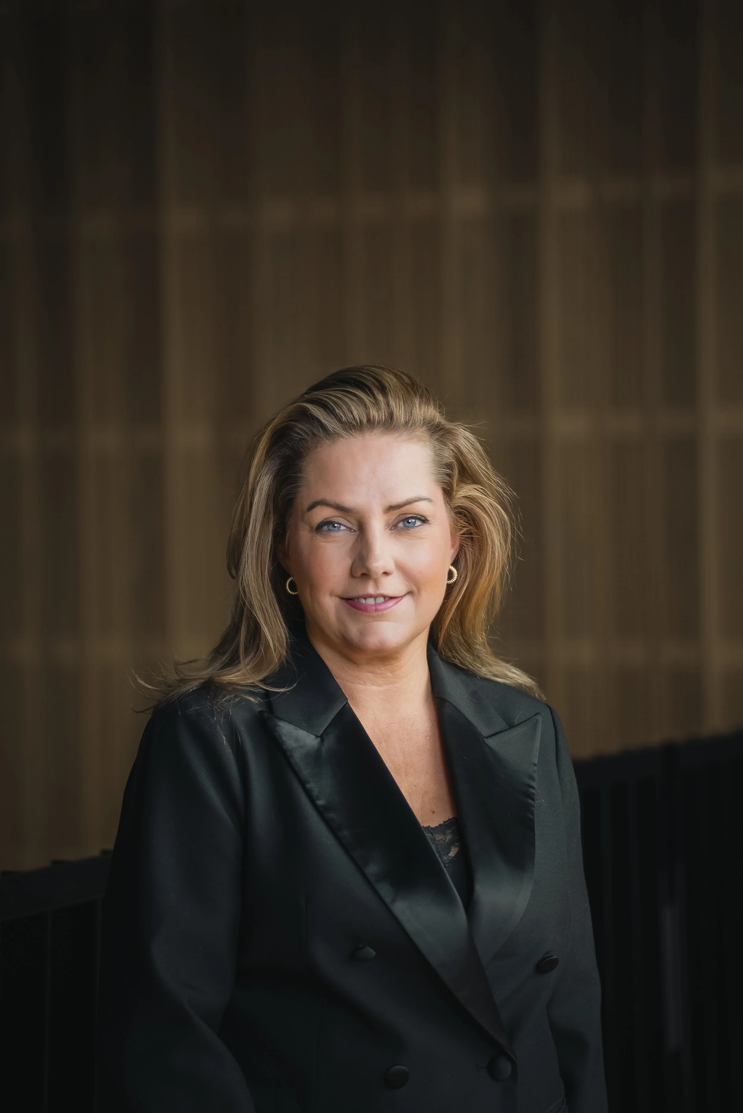 A middle-aged woman with light brown hair and blue eyes, smiling, wearing a black blazer, sitting indoors with a brown textured background.
