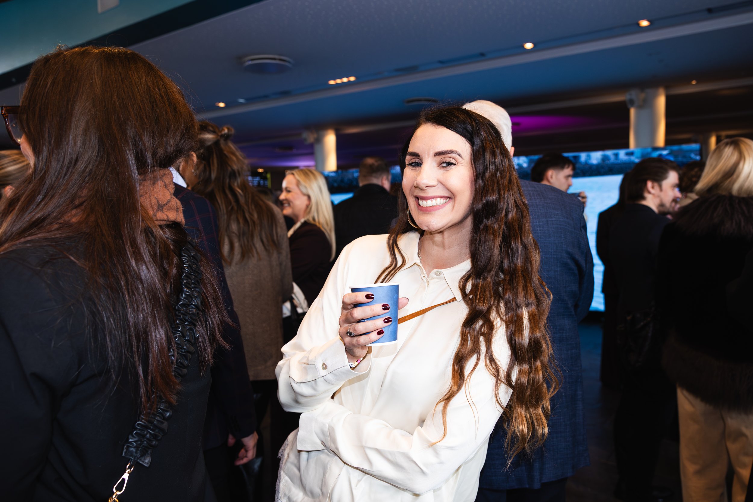 The image shows a smiling woman with long wavy brown hair holding a blue cup, engaged in conversation at a social gathering with multiple people in the background.
