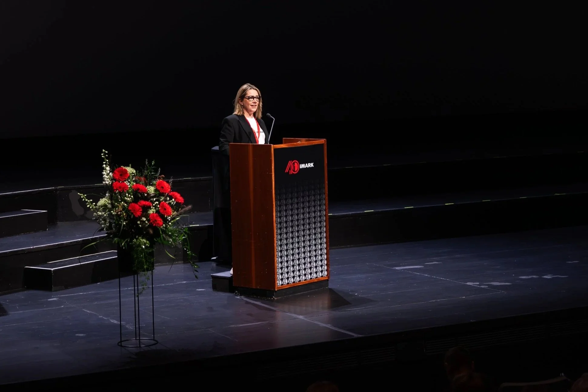 A woman speaking at a podium on a stage, with a large floral arrangement featuring red flowers nearby, in a dark auditorium setting.